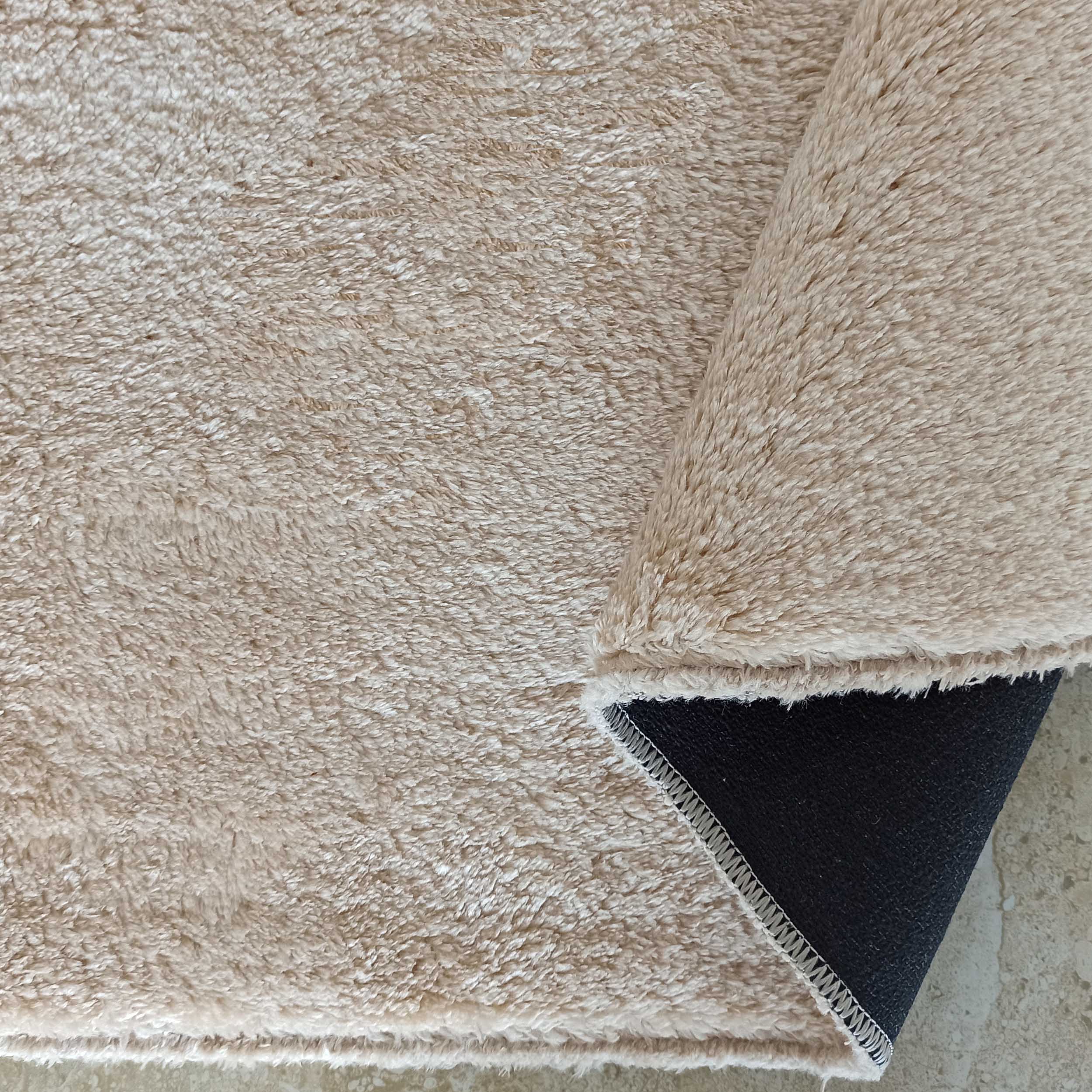 Tapis beige à poils longs et texture douce, un coin est plié révélant le dos noir antidérapant et les coutures blanches