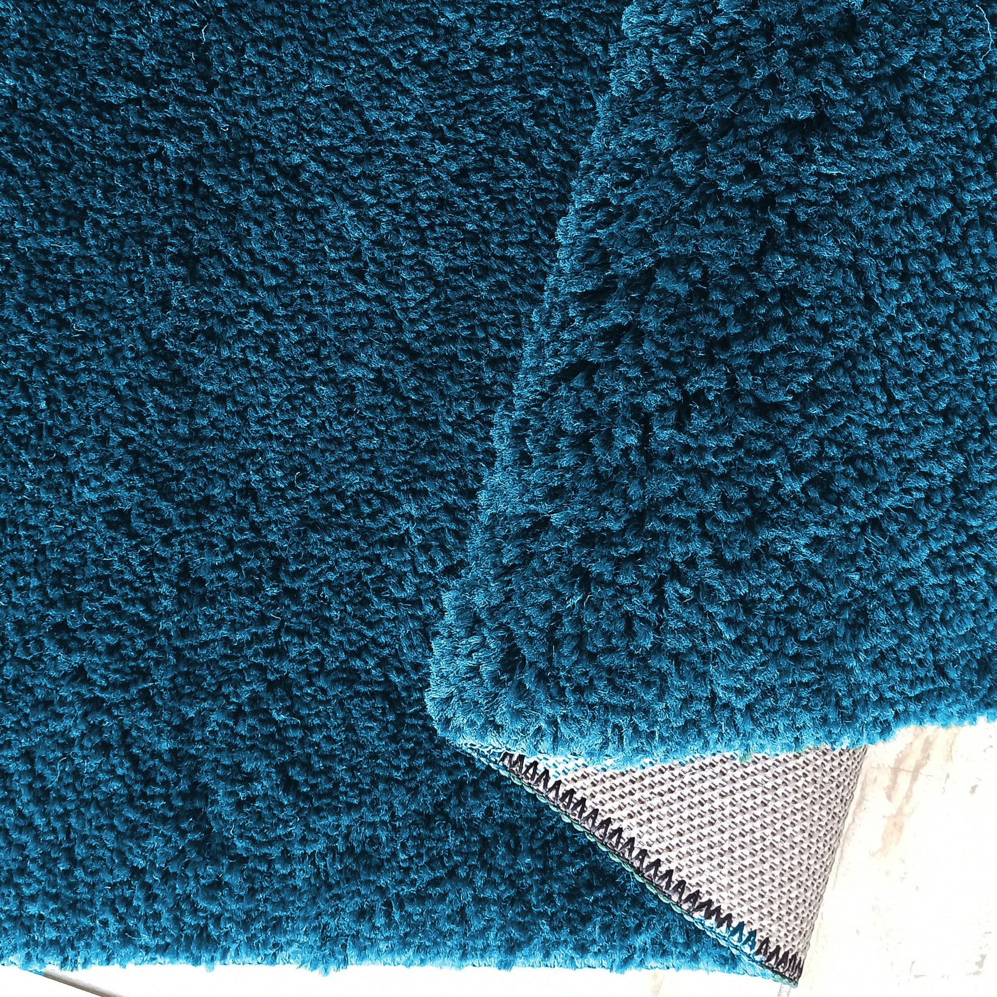 Tapis bleu sarcelle à poils longs, avec un coin replié montrant le revers gris quadrillé et une couture noire
