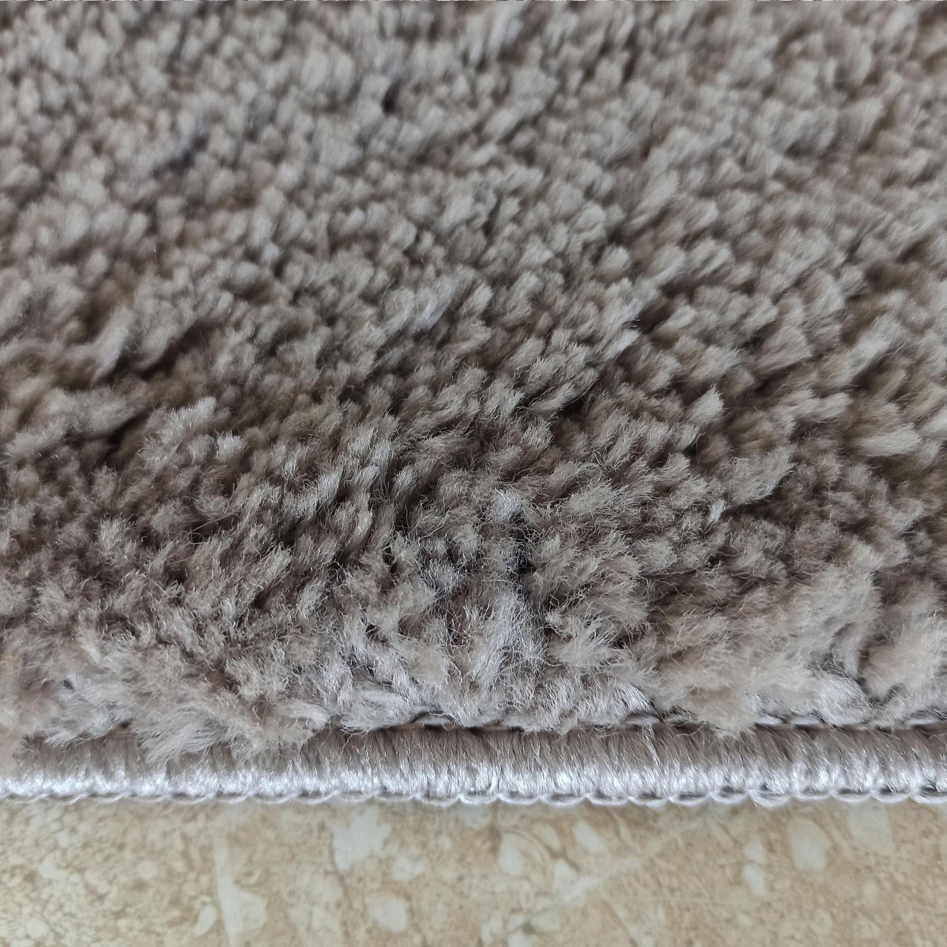 Tapis à poils longs et épais de couleur gris taupe, avec une bordure claire, posé sur un carrelage beige