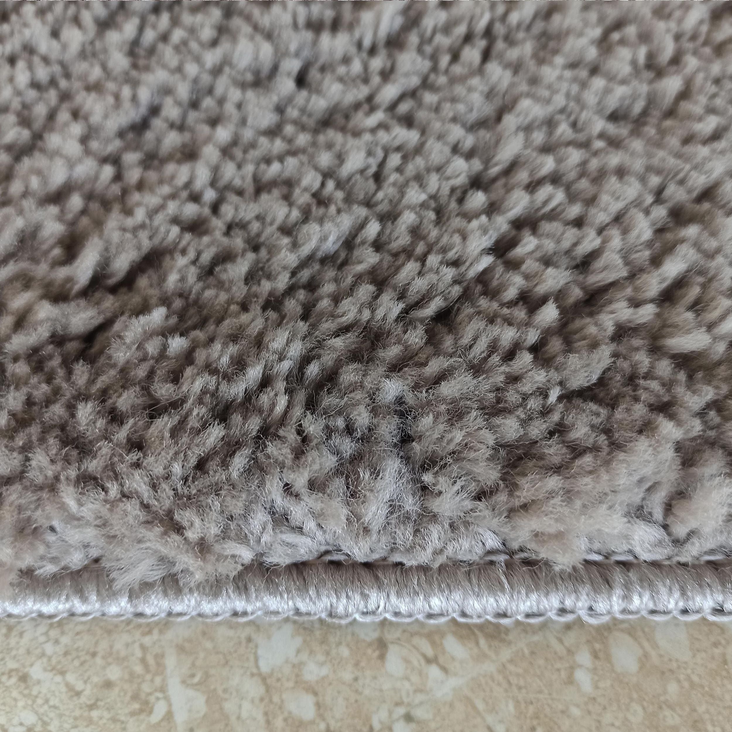 Tapis à poils longs et épais de couleur gris taupe, avec une bordure claire, posé sur un carrelage beige