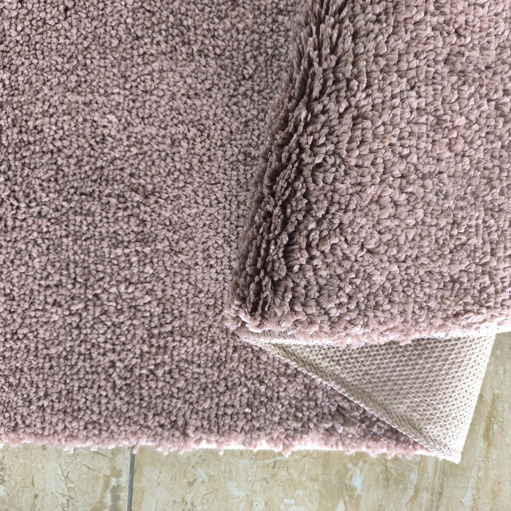 Tapis rose poudré à poils longs partiellement roulé, dévoilant son envers tissé et un sol carrelé beige clair