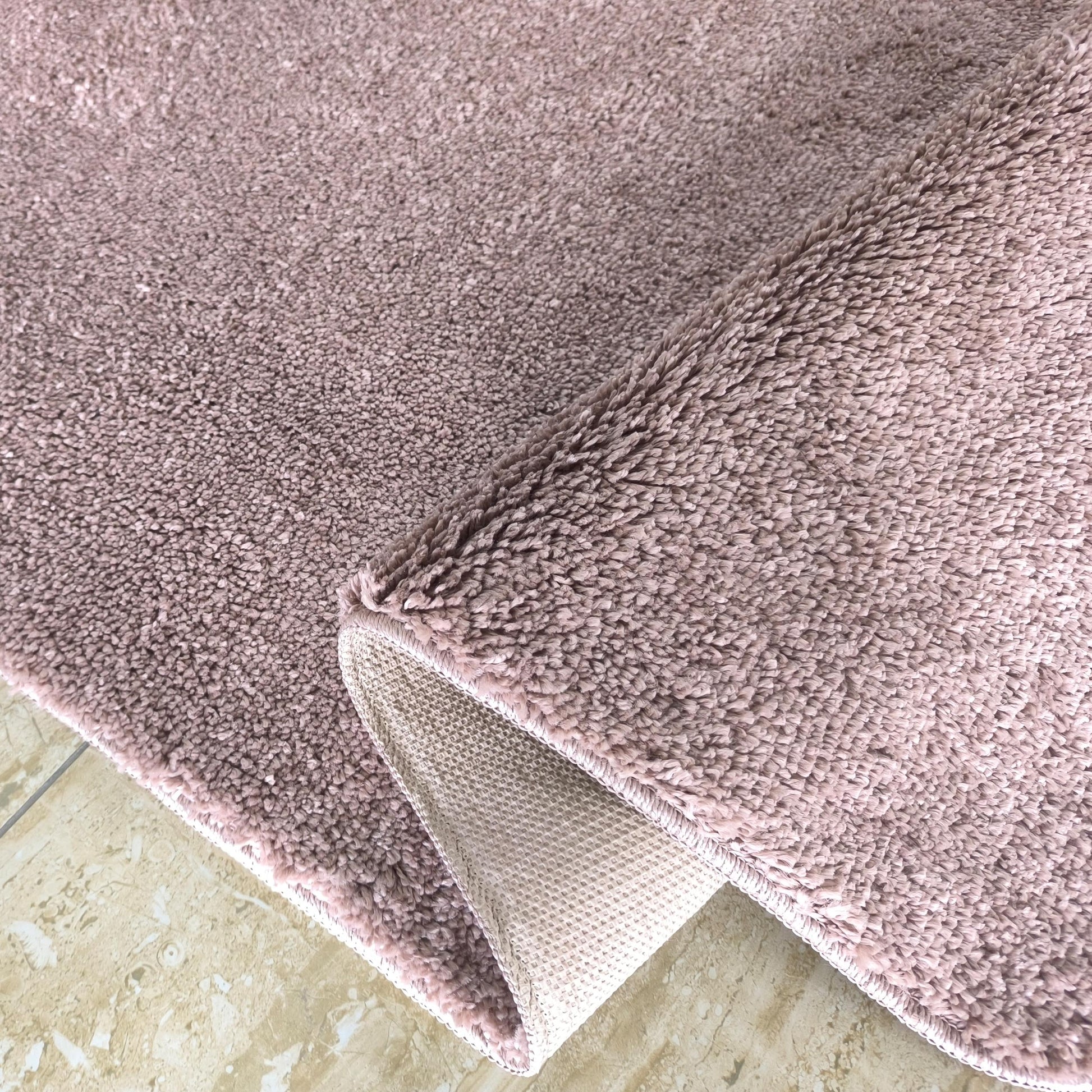 Tapis rose poudré à poils longs et moelleux, dont un coin est relevé, révélant son envers tissé sur un sol carrelé beige