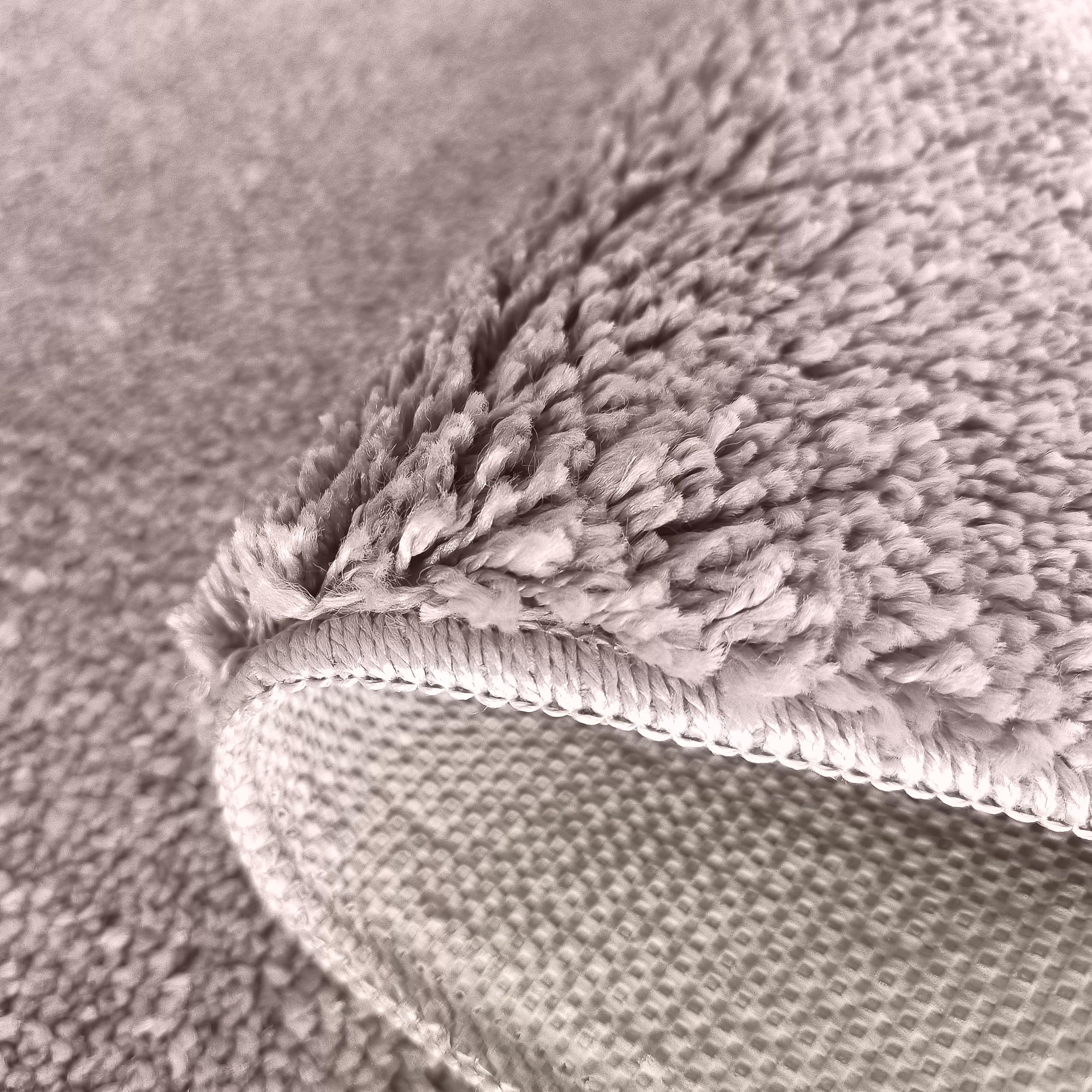 Gros plan d'un tapis taupe clair à la texture douce et moelleuse, plié pour révéler son revers quadrillé et la bordure cousue