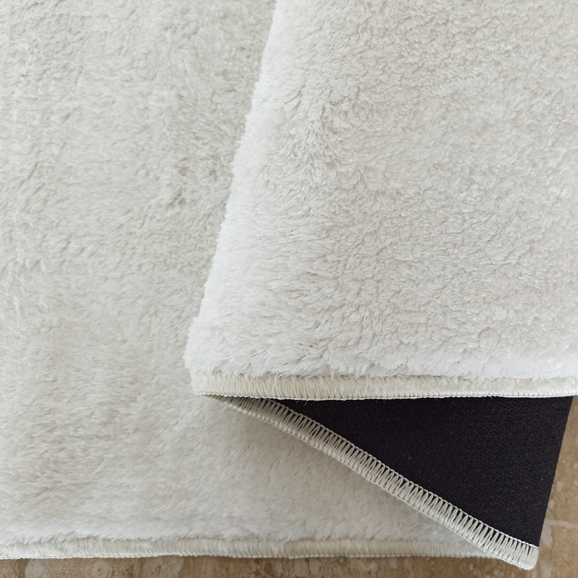 Tapis moelleux blanc cassé, un coin est replié montrant son dos antidérapant sombre et le sol carrelé beige