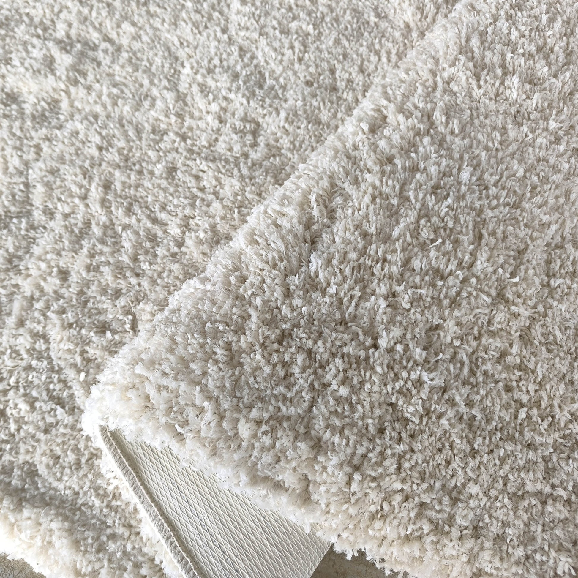 Tapis moelleux à poils longs de couleur blanc cassé, une partie est repliée laissant apparaître l'envers texturé