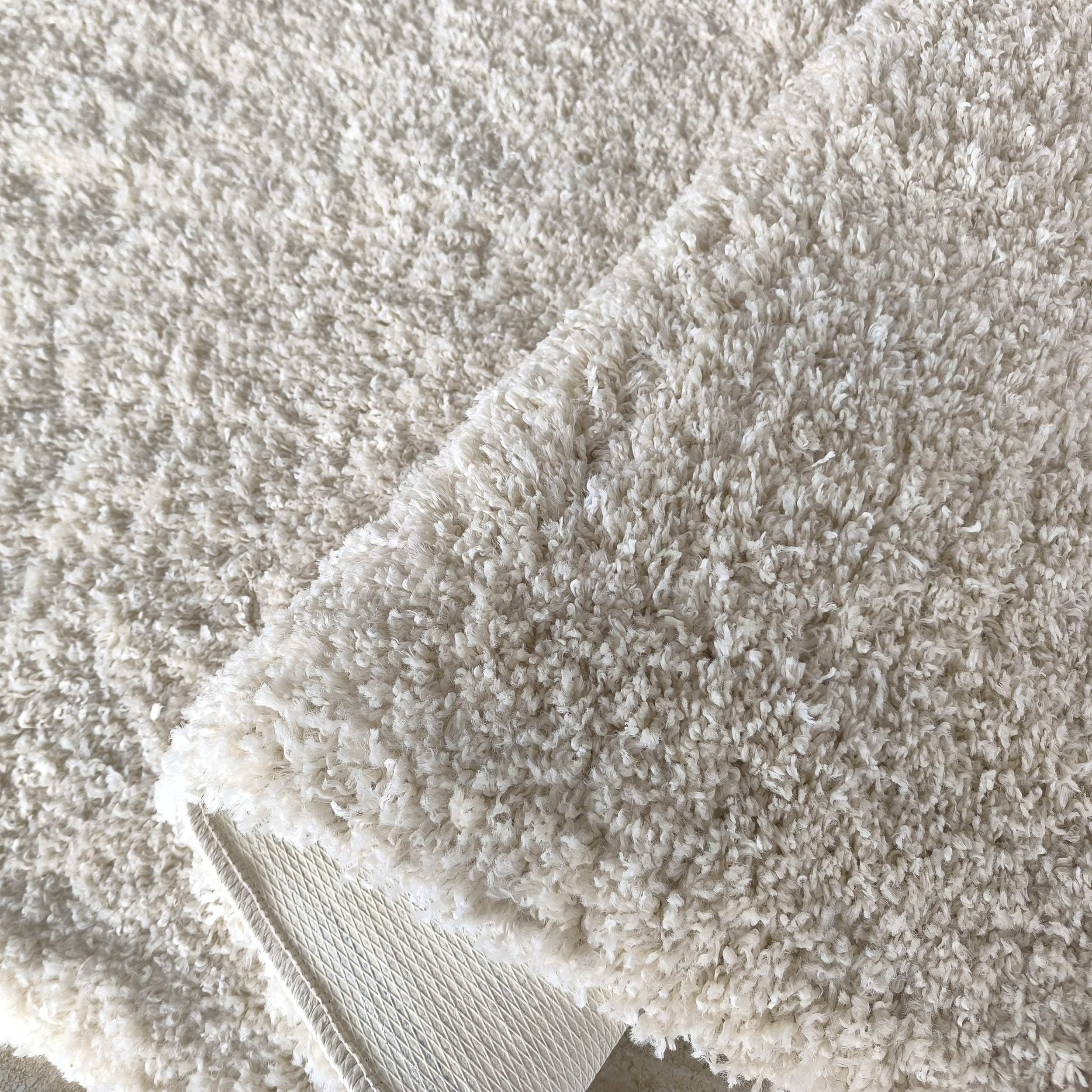 Tapis moelleux à poils longs de couleur blanc cassé, une partie est repliée laissant apparaître l'envers texturé