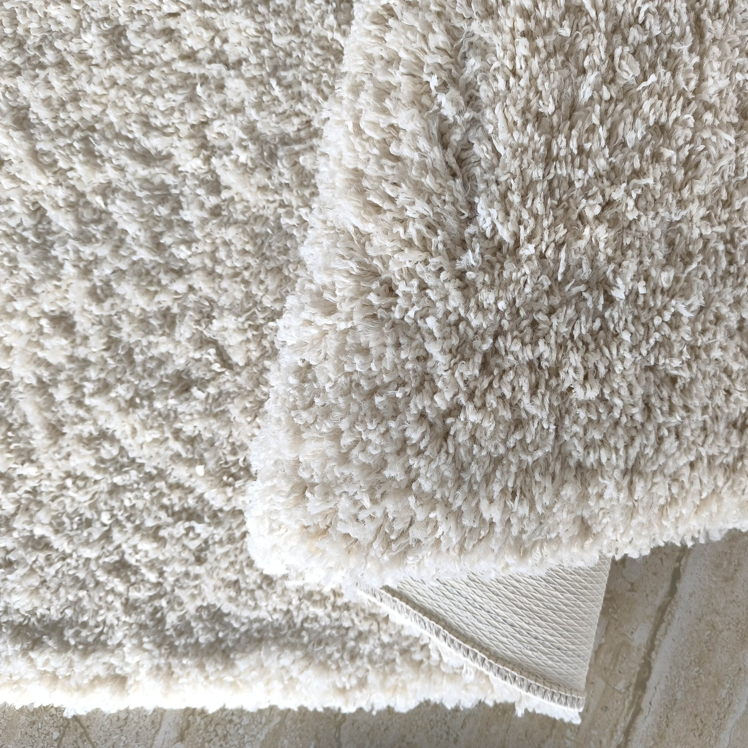 Tapis moelleux et blanc cassé à longs poils bouclés, partiellement replié, laissant apparaître son envers sur un sol beige