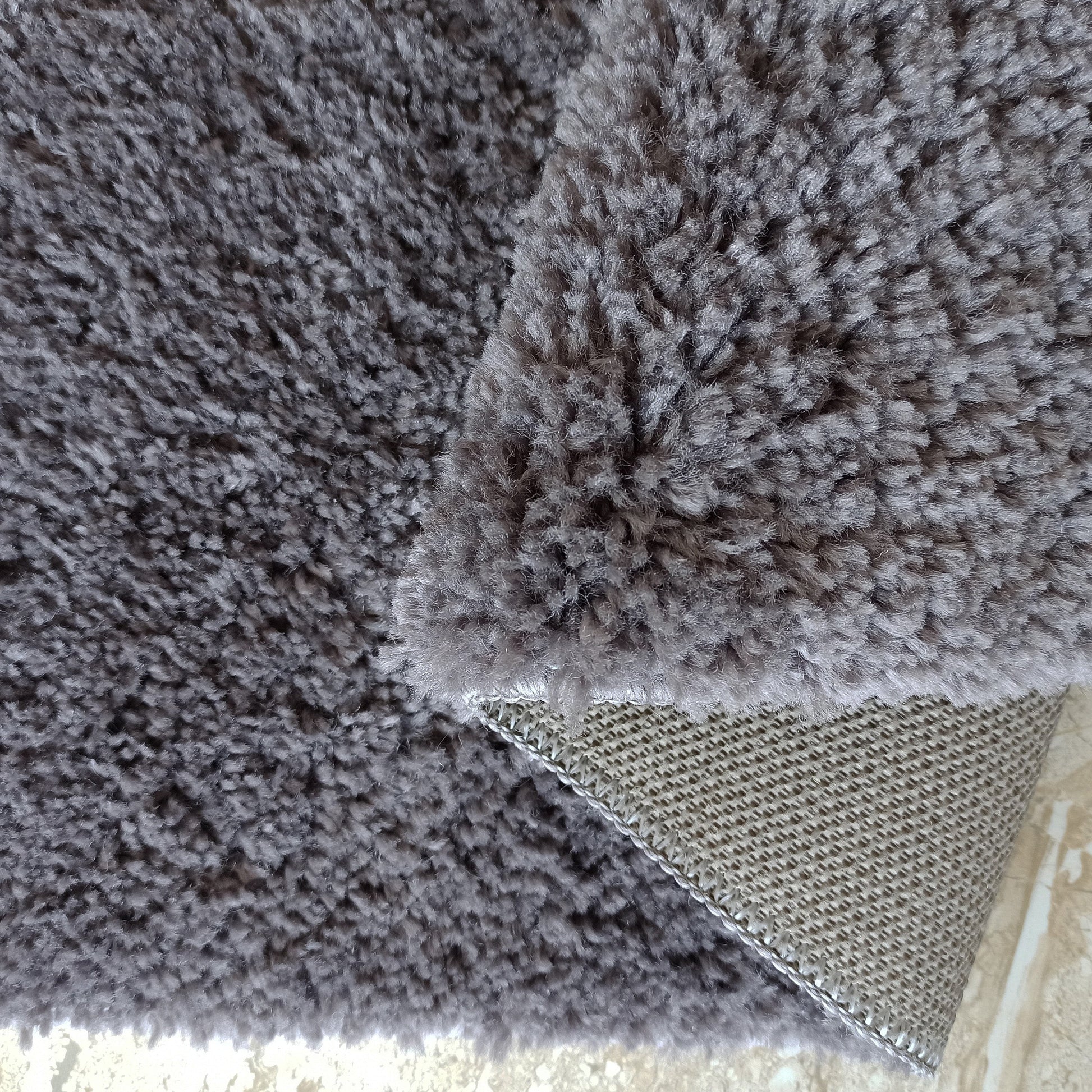 Gros plan sur un tapis gris anthracite à longs poils, un coin soulevé dévoile son envers tissé à motif quadrillé beige