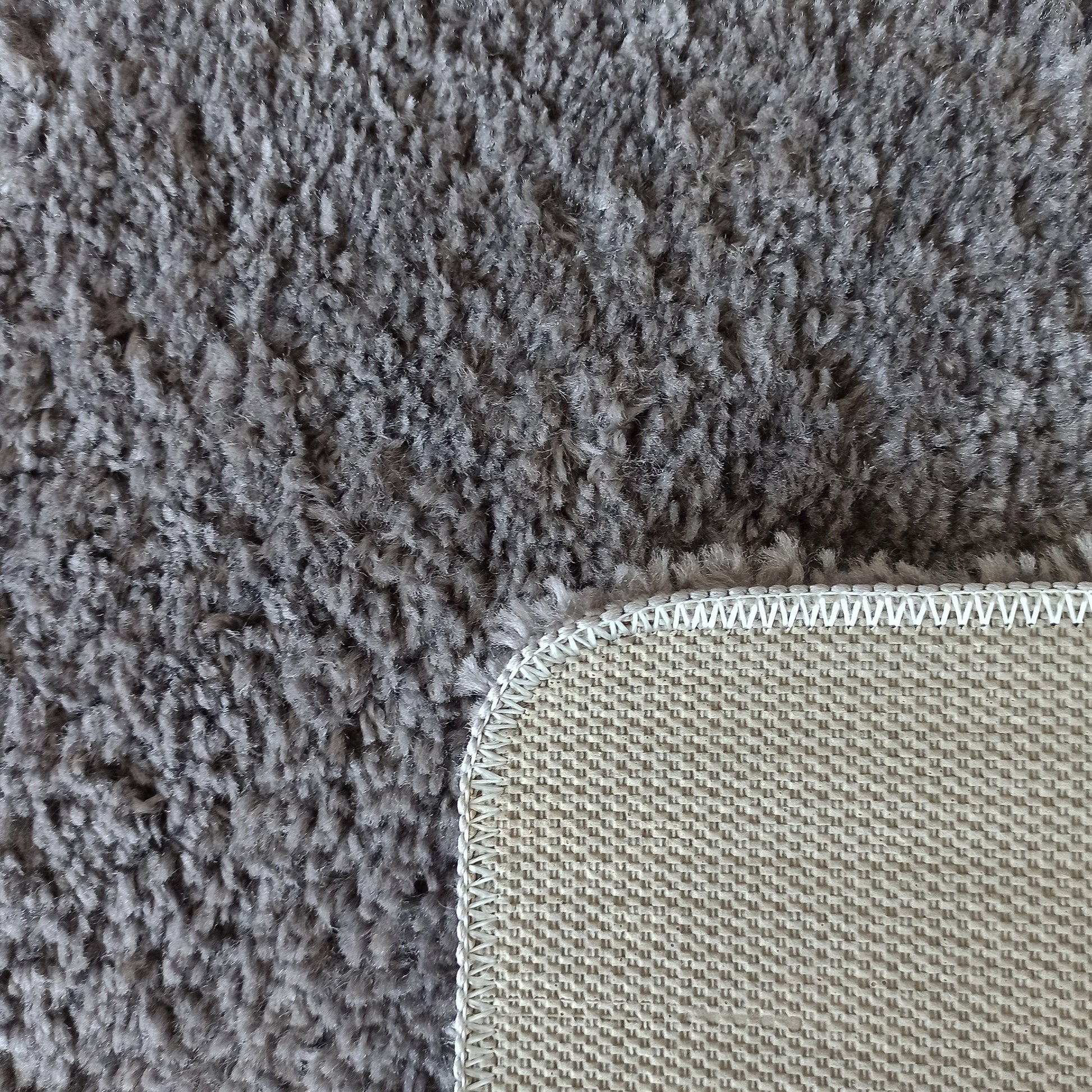 Tapis gris à longs poils. Son coin relevé montre le dessous beige clair en tissu tissé et la bordure blanche surpiquée