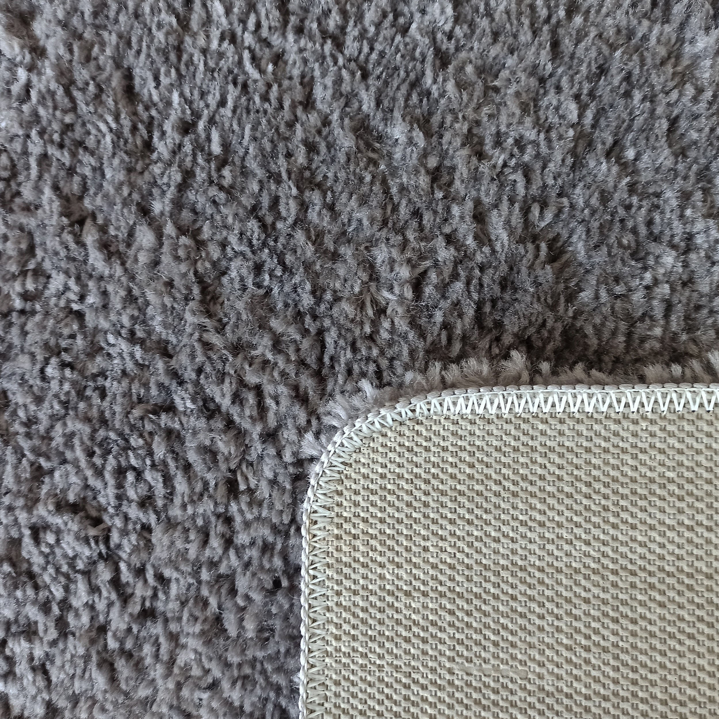 Tapis gris à longs poils. Son coin relevé montre le dessous beige clair en tissu tissé et la bordure blanche surpiquée