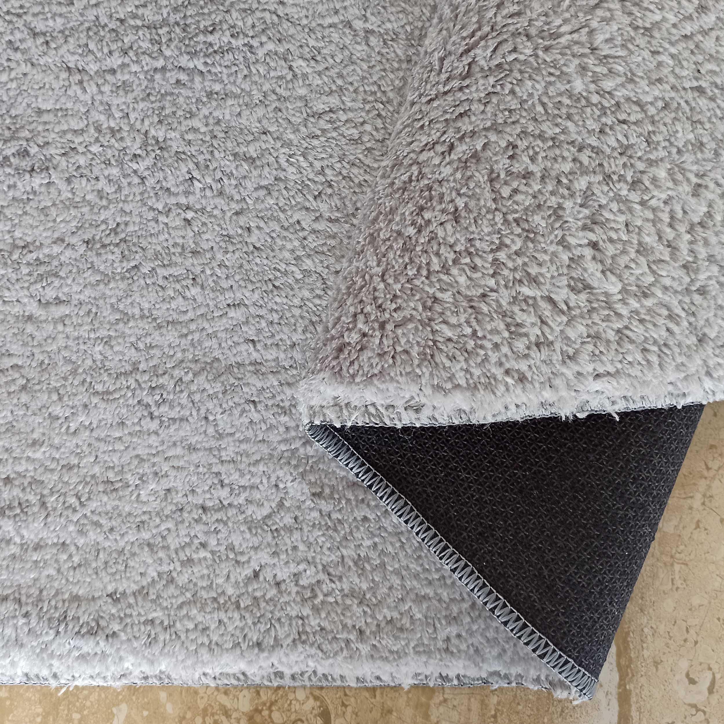 Gros plan sur un tapis gris clair à poils longs et doux, un coin est relevé exposant l'envers noir antidérapant et le sol carrelé beige