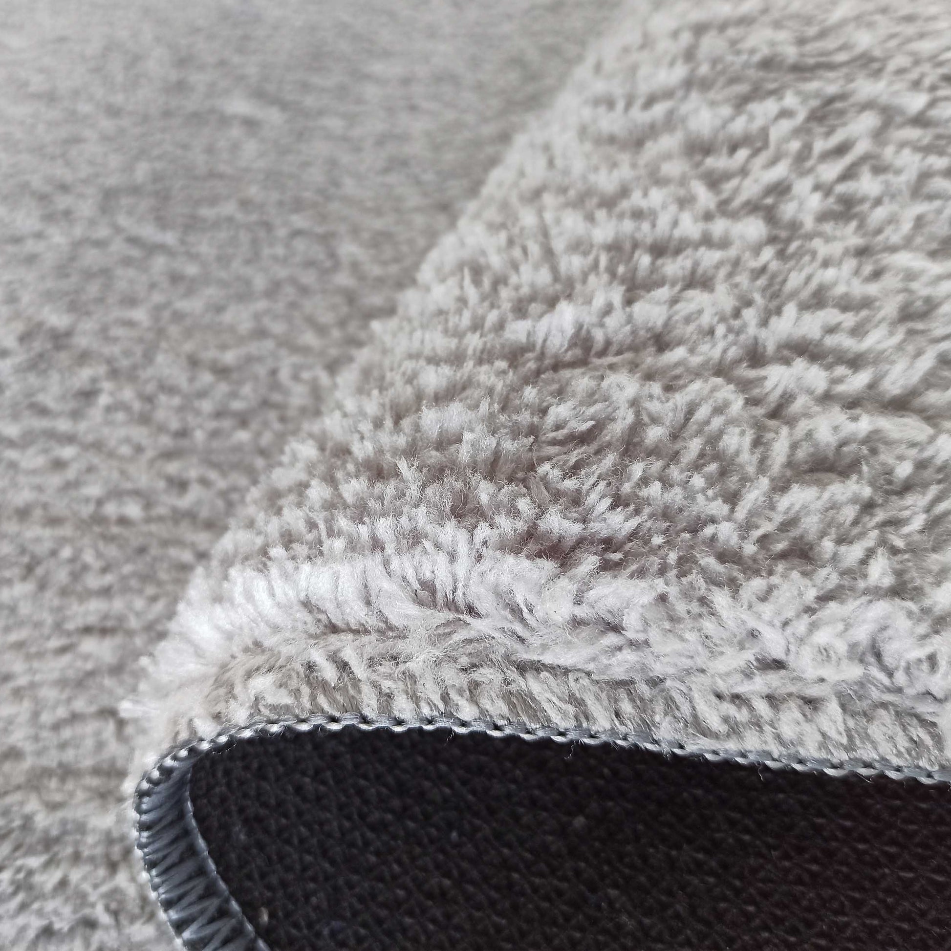 Gros plan sur un tapis gris clair à longs poils doux, avec son envers foncé texturé et une bordure cousue apparente