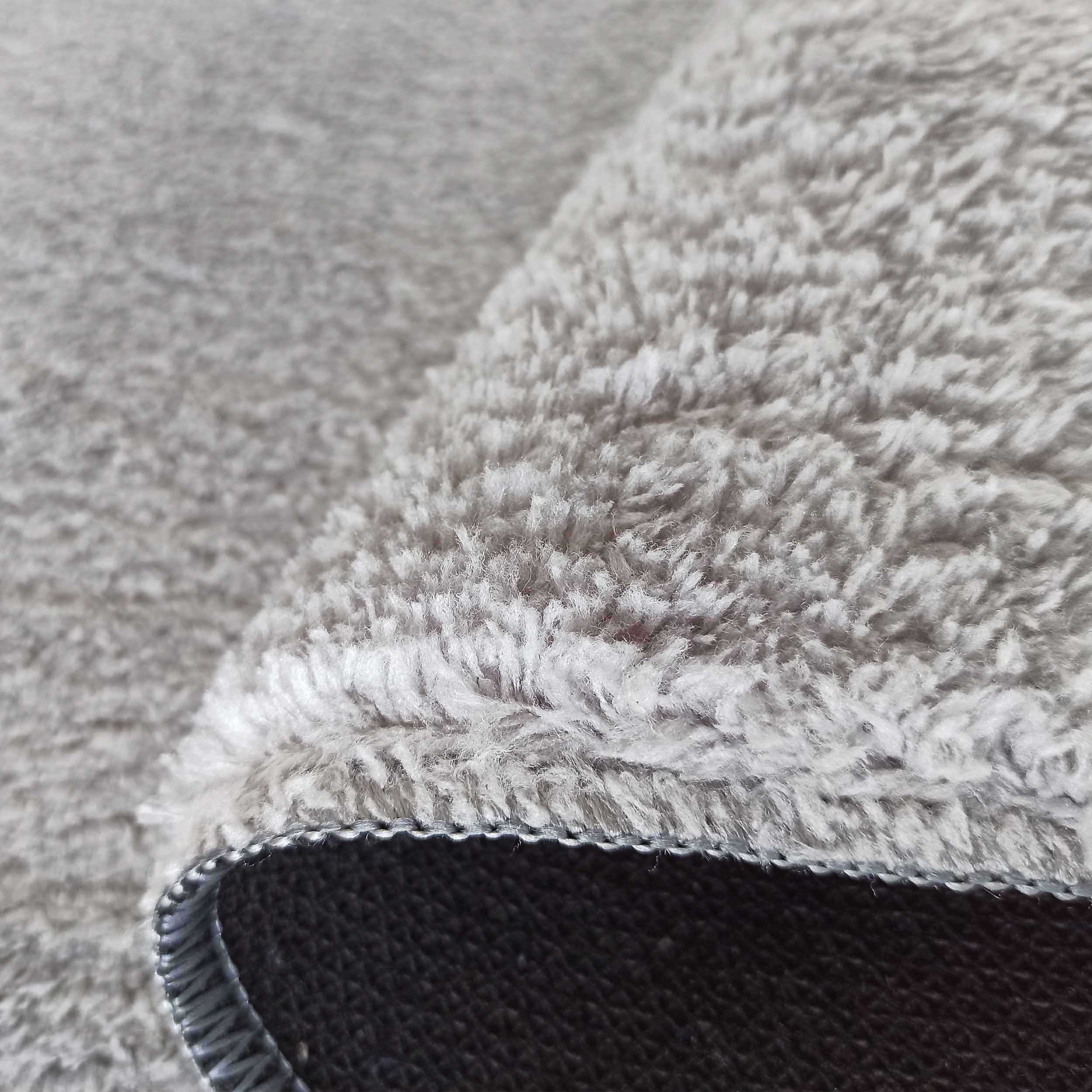 Gros plan sur un tapis gris clair à longs poils doux, avec son envers foncé texturé et une bordure cousue apparente