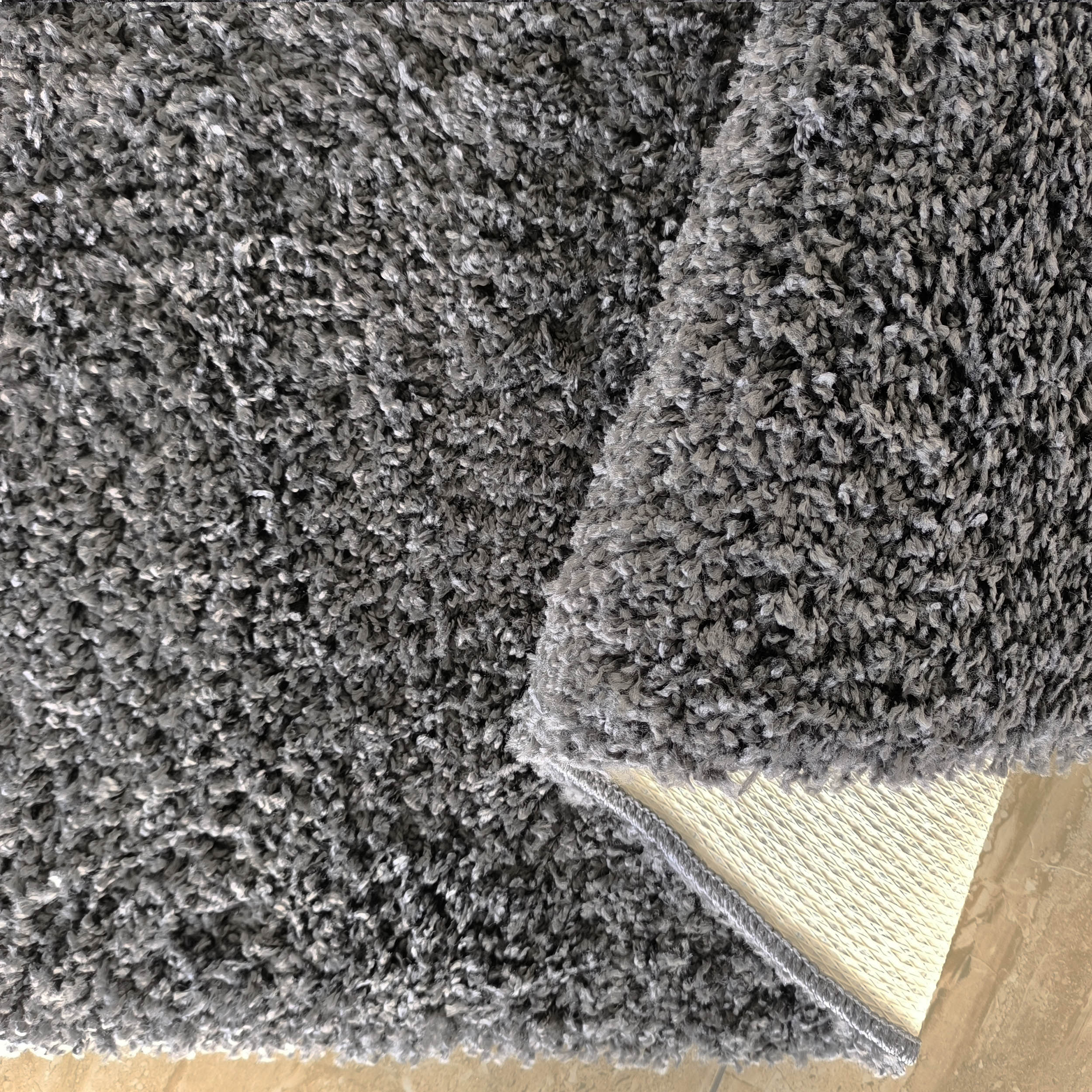 Tapis gris anthracite à longs poils mélangés, partiellement replié montrant son envers texturé beige sur un carrelage clair