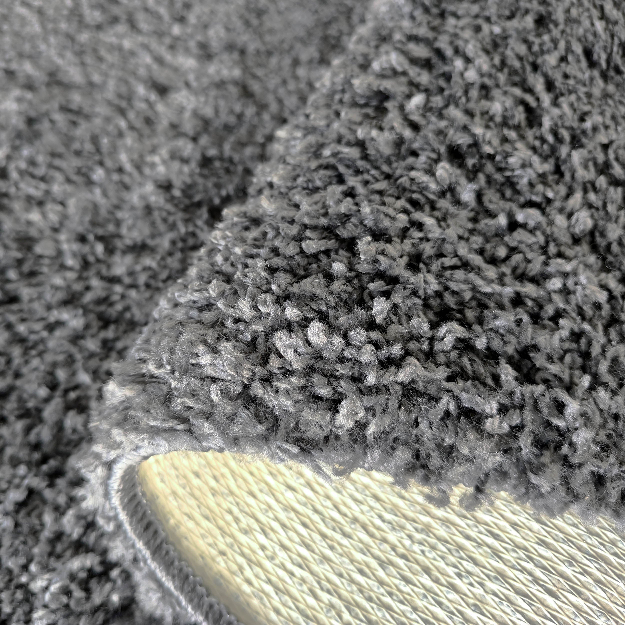 Tapis gris à poils longs et denses, montrant sa texture douce. Le tapis est relevé, révélant son envers tissé clair au motif quadrillé