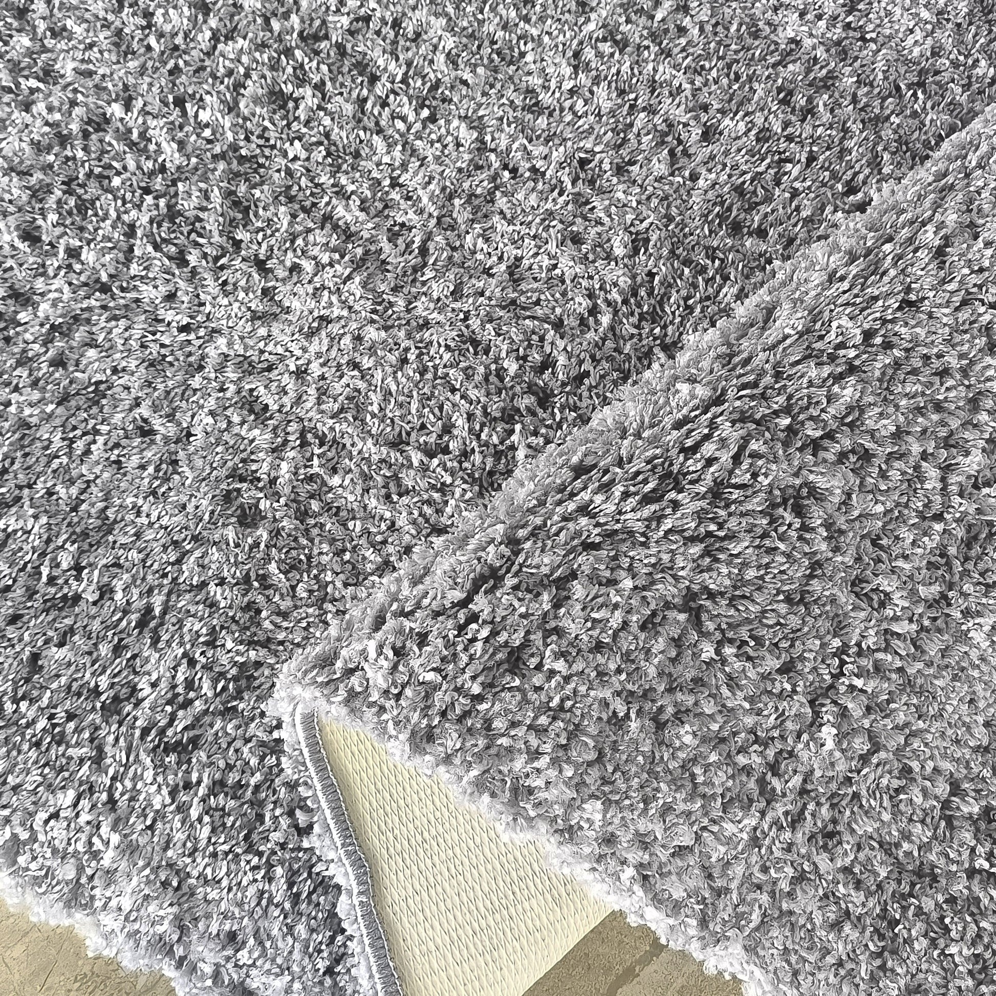Gros plan sur un tapis gris moelleux à la texture bouclée, avec un coin relevé dévoilant son envers beige quadrillé