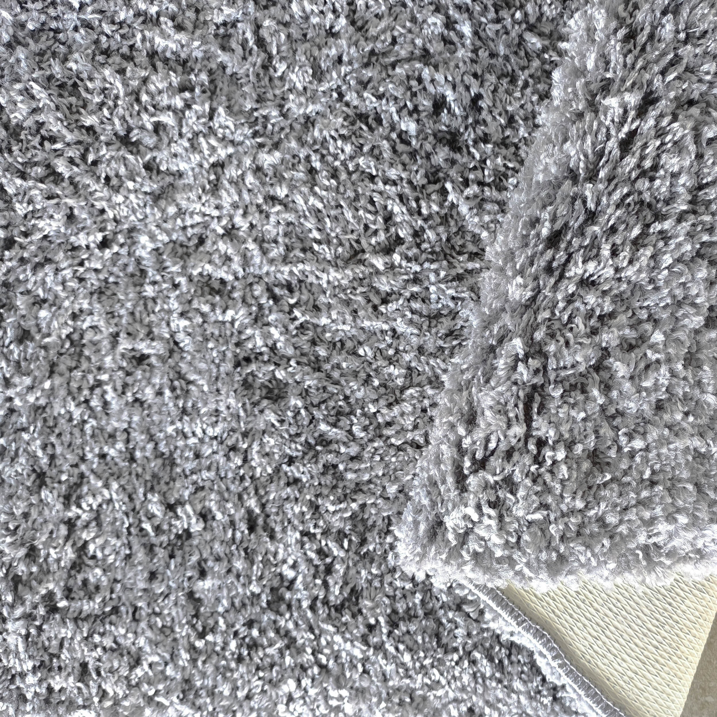 Tapis moelleux aux teintes de gris et argent, avec des fibres épaisses. Une partie est relevée, révélant le dessous beige texturé