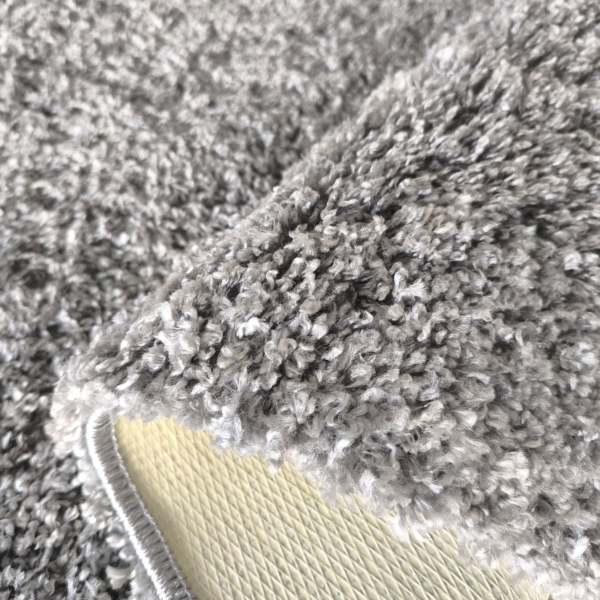 Tapis moelleux gris foncé et clair, avec un coin relevé montrant son envers crème à motifs de losanges et sa bordure cousue