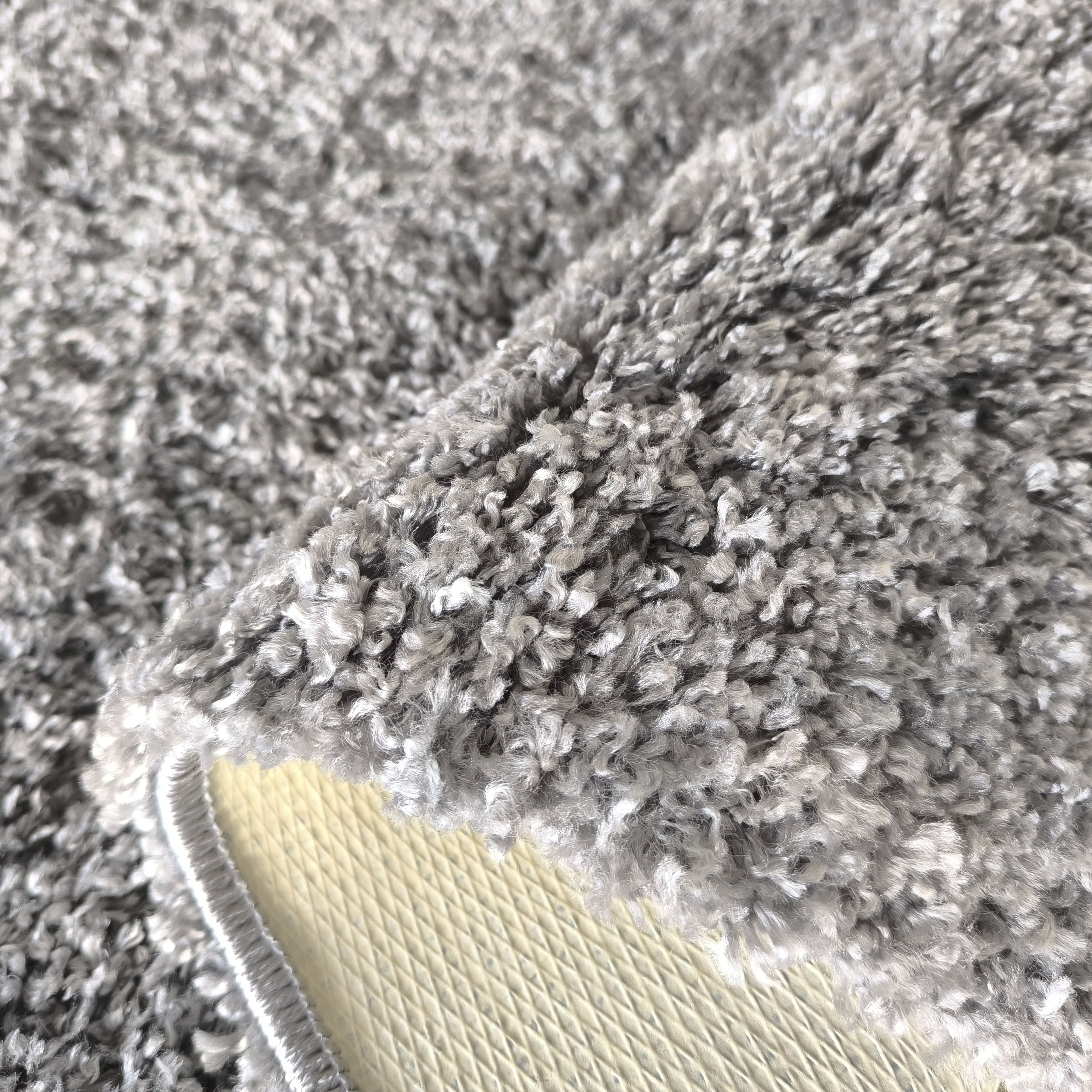 Tapis moelleux gris foncé et clair, avec un coin relevé montrant son envers crème à motifs de losanges et sa bordure cousue
