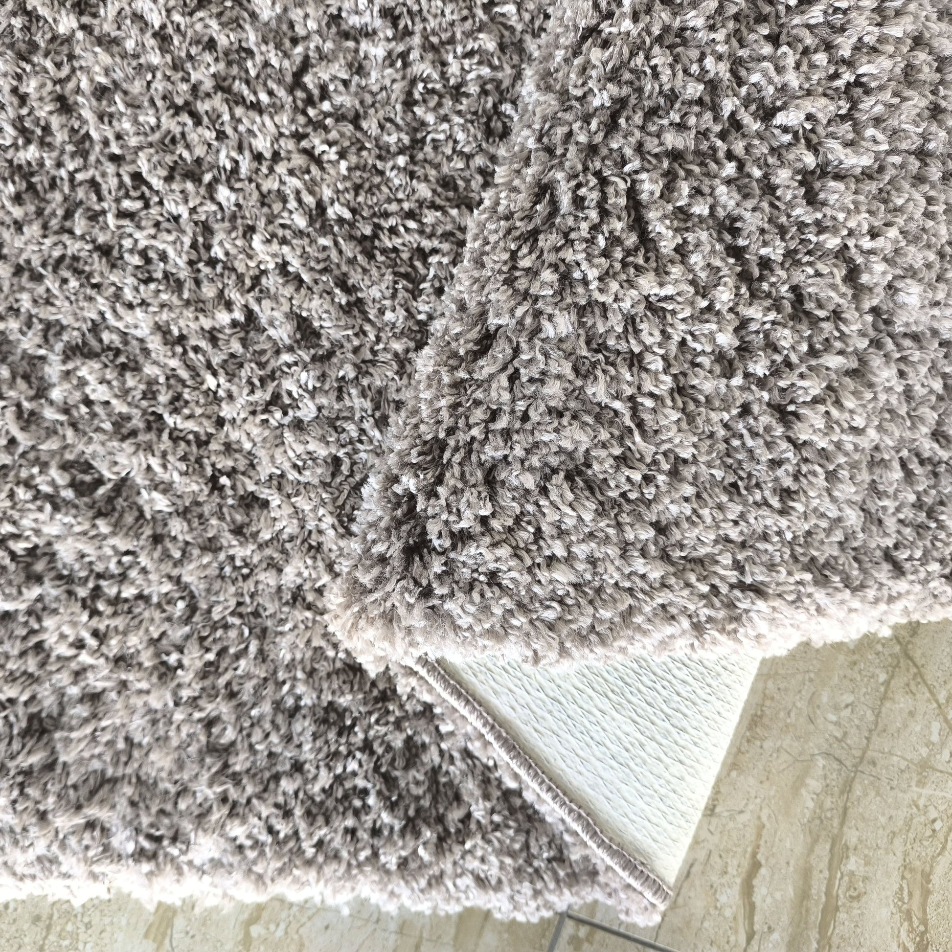 Tapis épais à poils longs et bouclés gris-beige clair chiné, dont un coin est relevé sur un sol carrelé beige