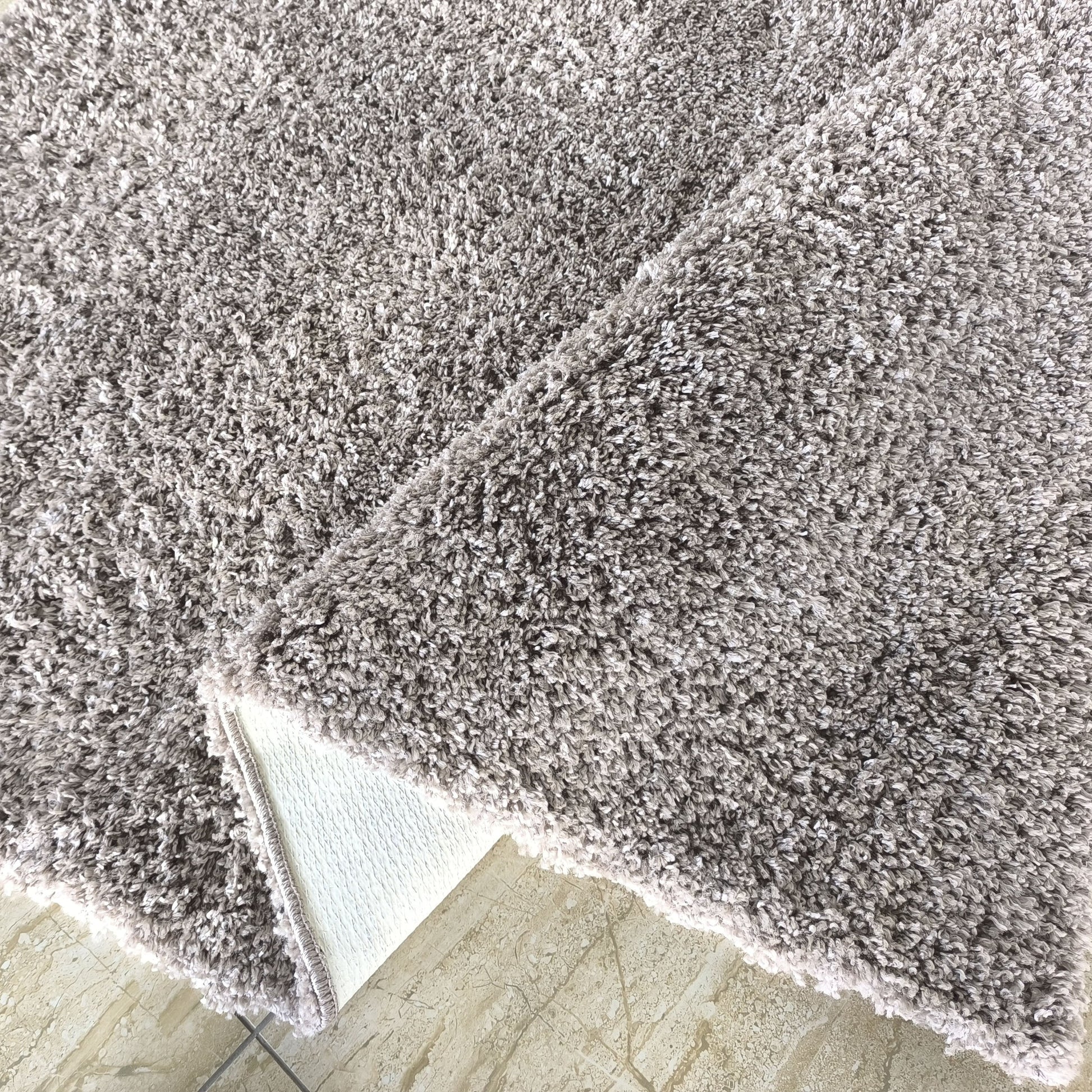 Tapis gris chiné à poils longs et moelleux, partiellement plié, révélant son envers blanc sur un carrelage beige