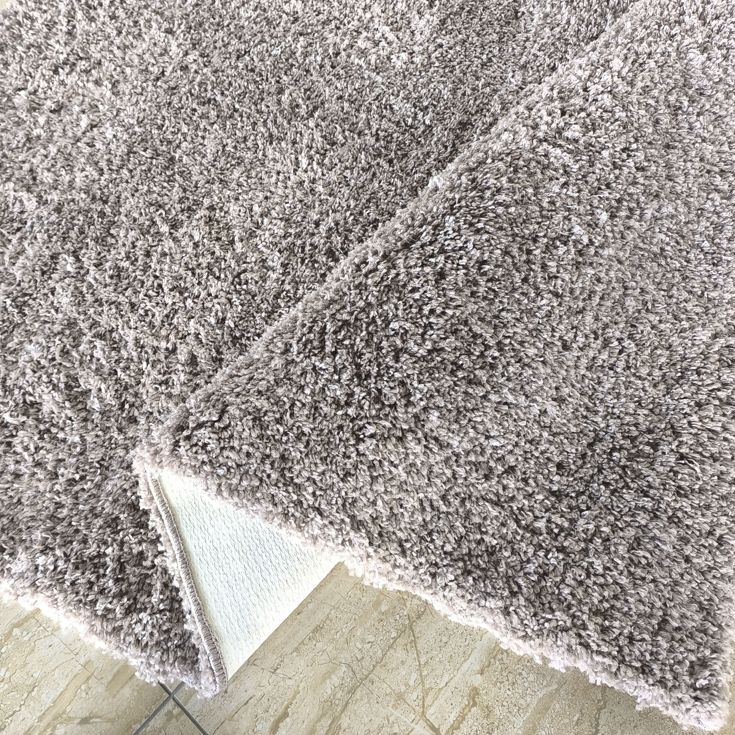 Tapis gris chiné à poils longs et moelleux, partiellement plié, révélant son envers blanc sur un carrelage beige