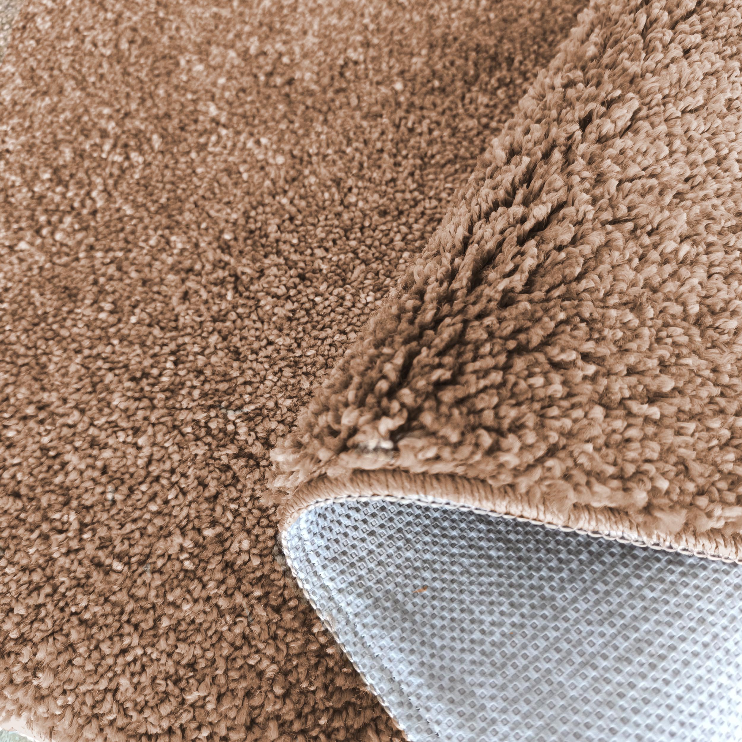 Tapis couleur taupe à texture douce et veloutée, avec un coin replié montrant le dessous antidérapant gris à motif losanges