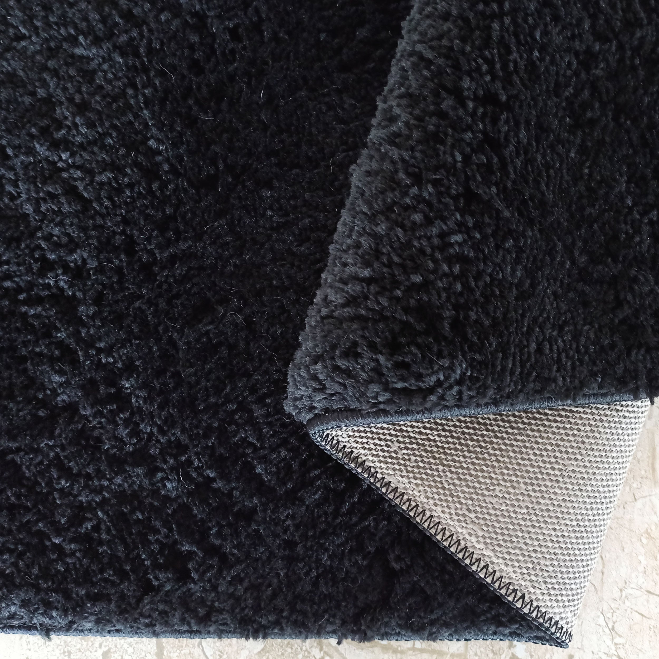 Tapis noir à poils longs et doux. Un coin est replié, montrant l'envers clair avec un motif tissé en petits carreaux et des coutures foncées