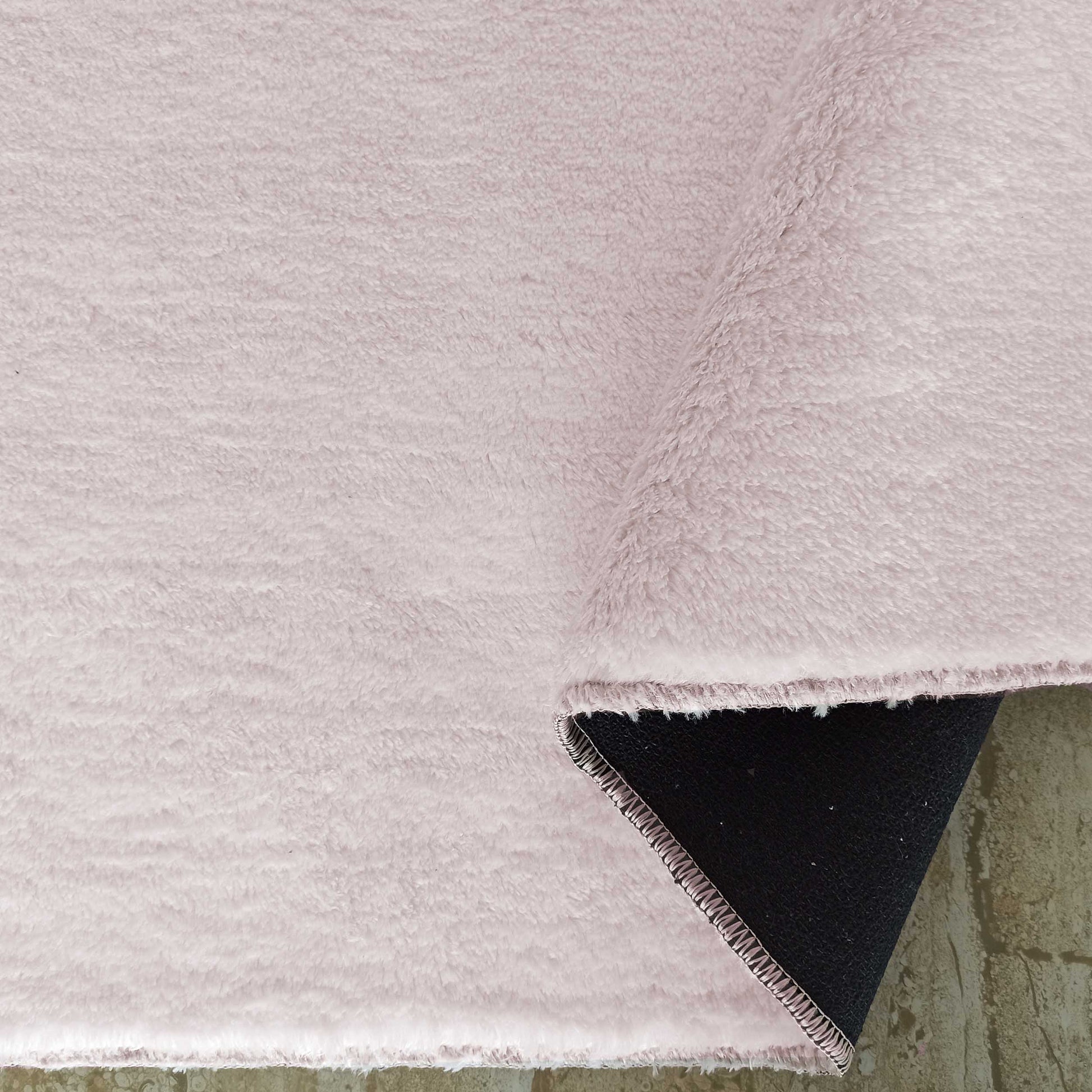 Tapis rose pâle à la texture douce et poilue, partiellement enroulé révélant son envers noir sur un sol clair texturé