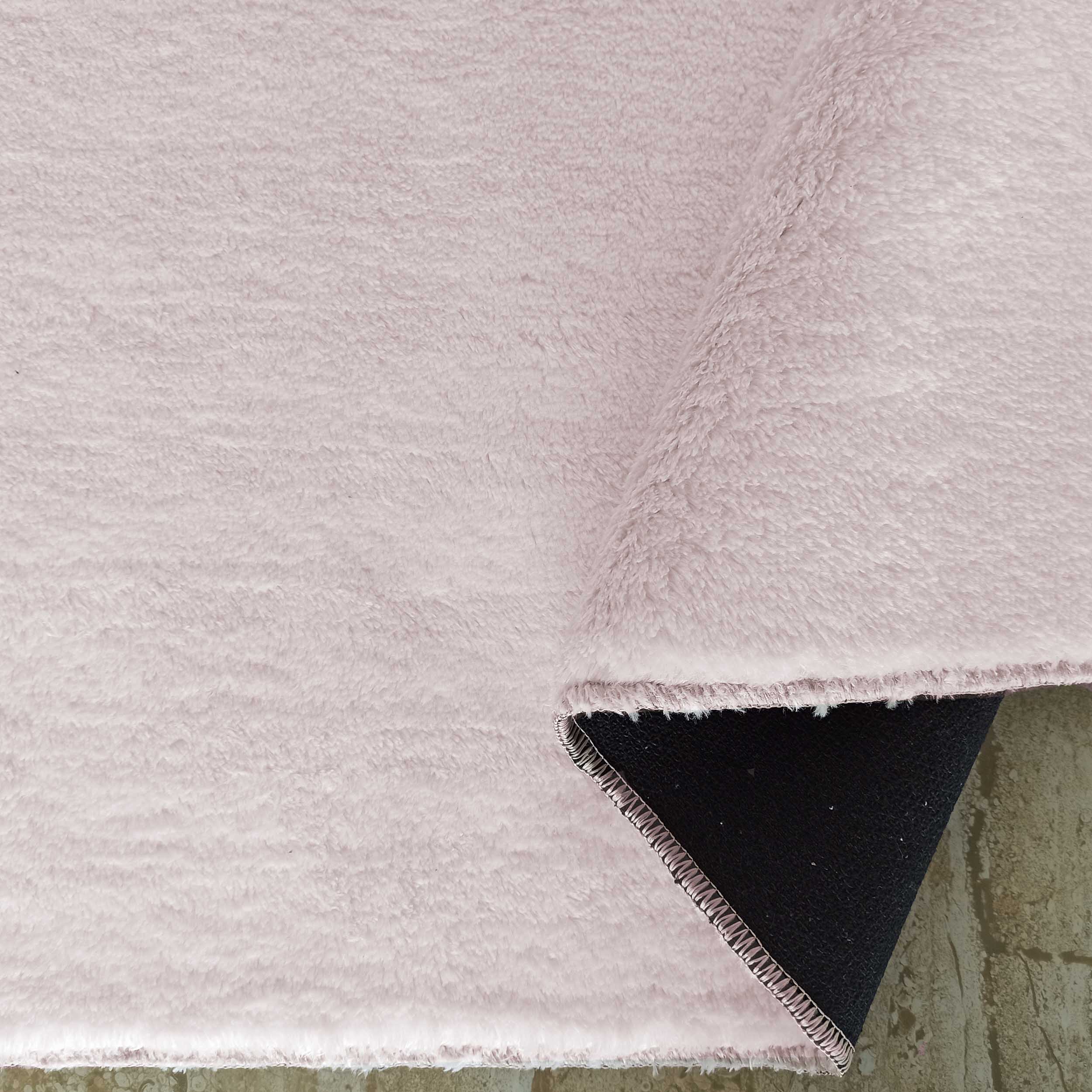 Tapis rose pâle à la texture douce et poilue, partiellement enroulé révélant son envers noir sur un sol clair texturé
