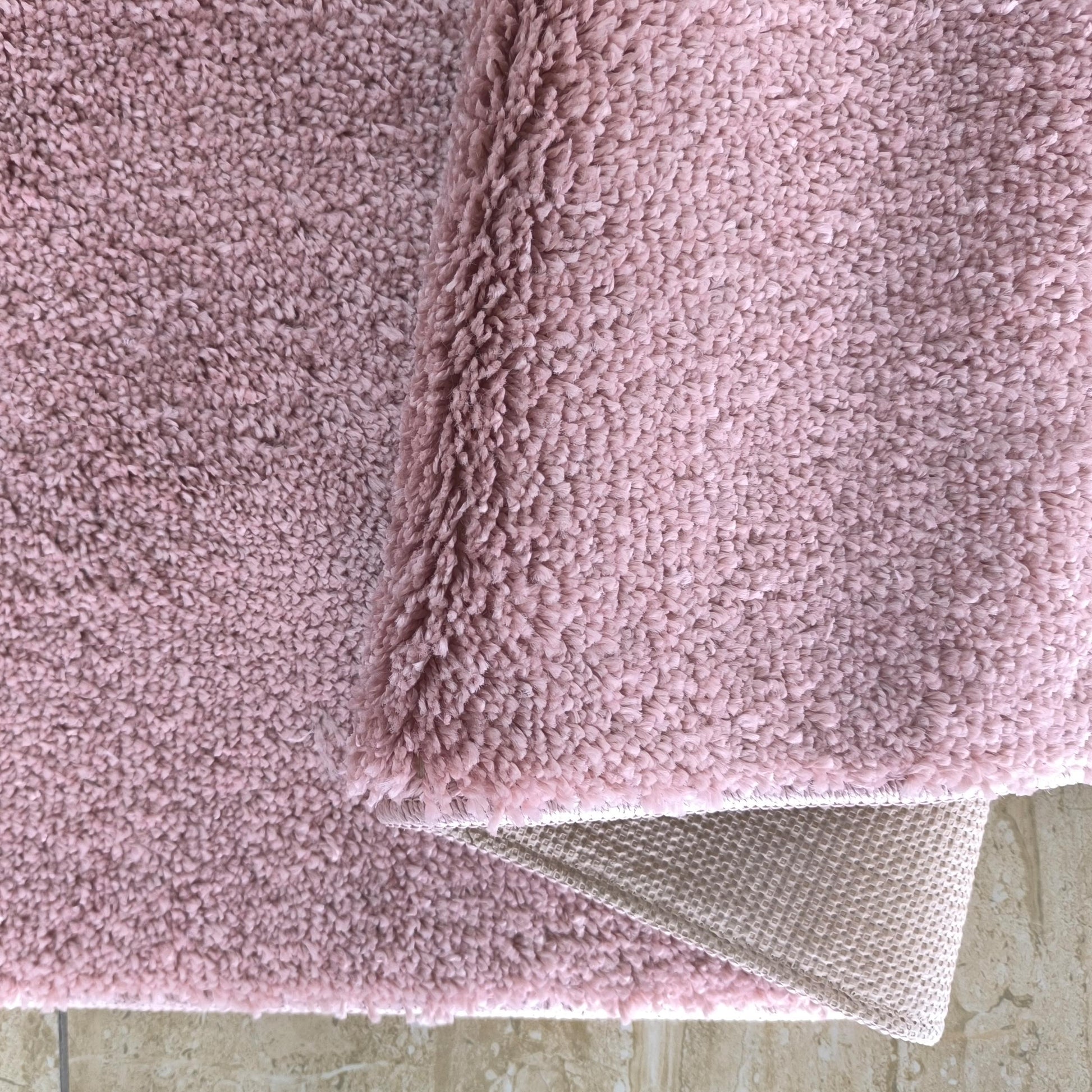 Tapis rose à la texture bouclée et douce, partiellement plié sur un carrelage clair, laissant voir son envers clair et tissé