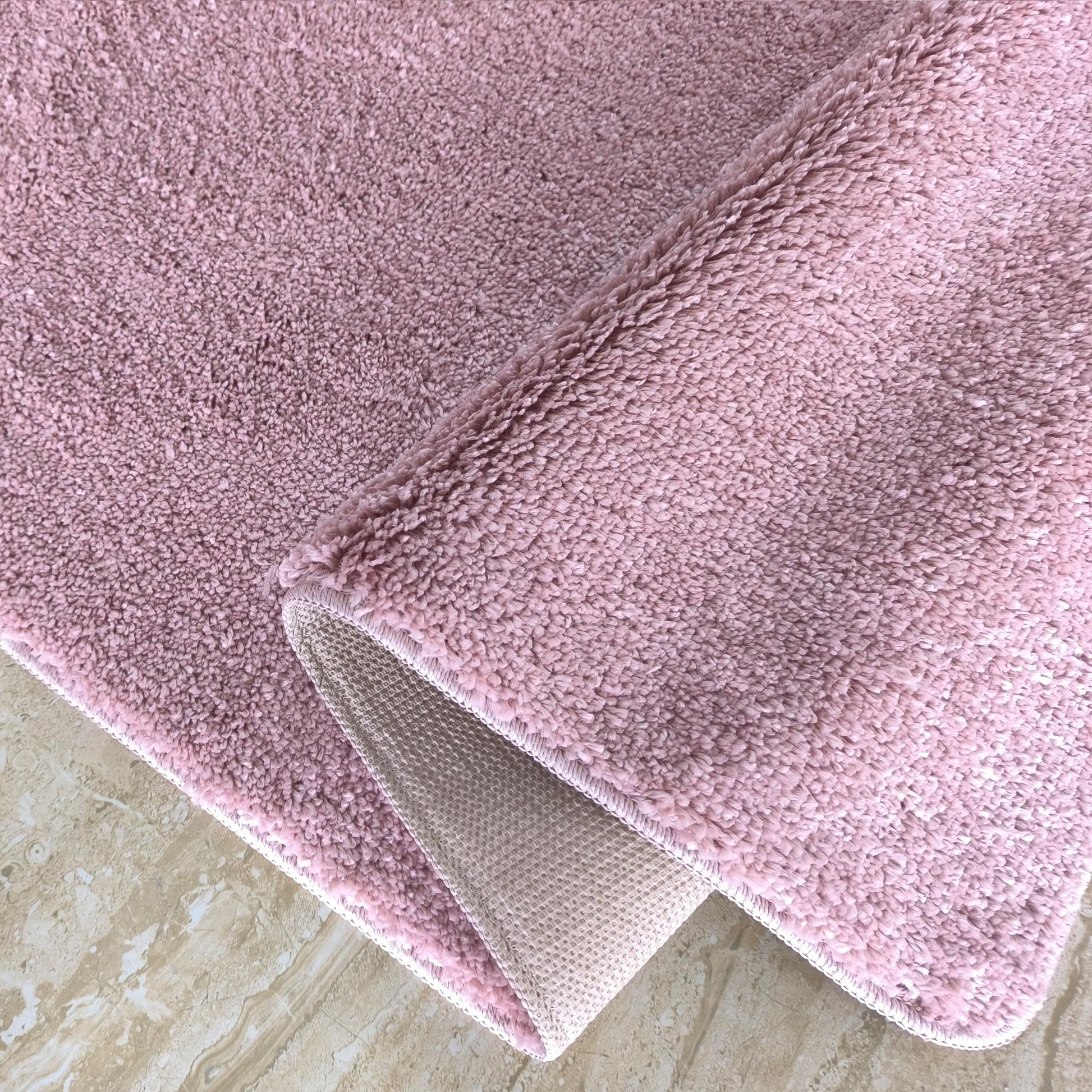 Tapis rose clair à texture douce et moelleuse, partiellement replié, révélant son envers antidérapant sur un sol marbré
