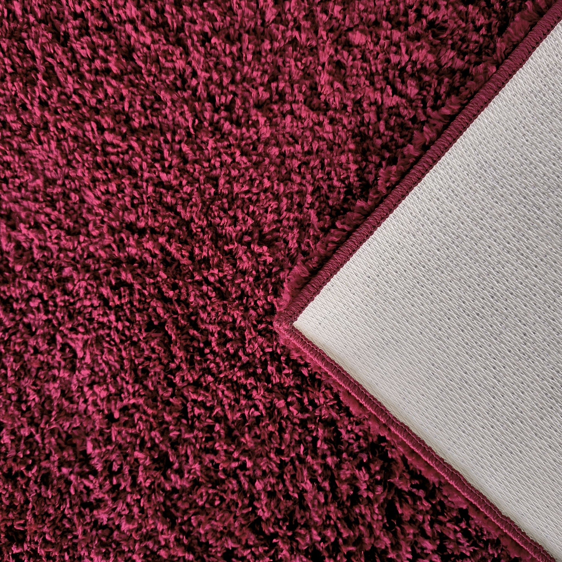 Tapis à poils longs couleur magenta foncé, dont un coin est relevé pour montrer l'envers texturé gris clair et la bordure cousue assortie