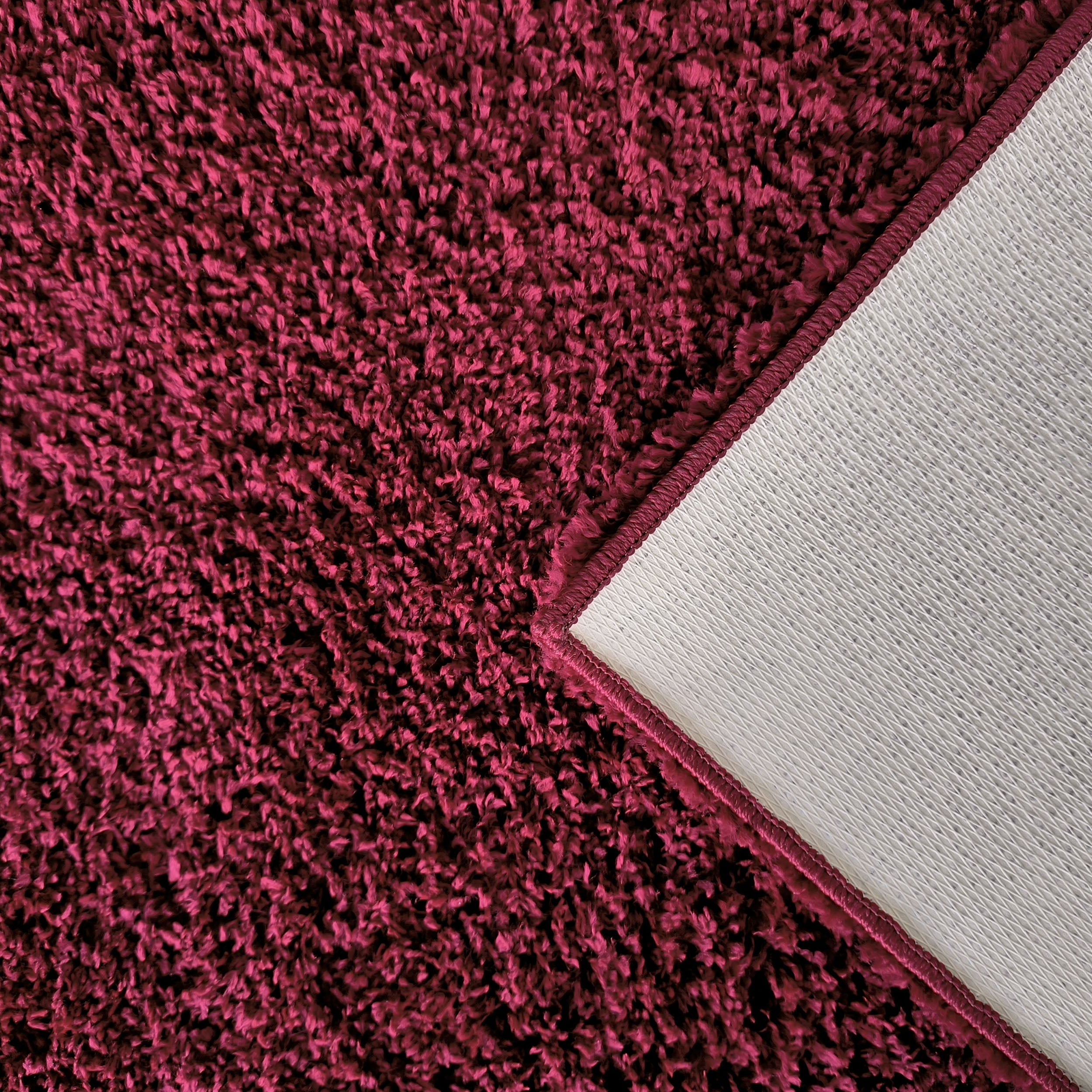 Tapis à poils longs couleur magenta foncé, dont un coin est relevé pour montrer l'envers texturé gris clair et la bordure cousue assortie