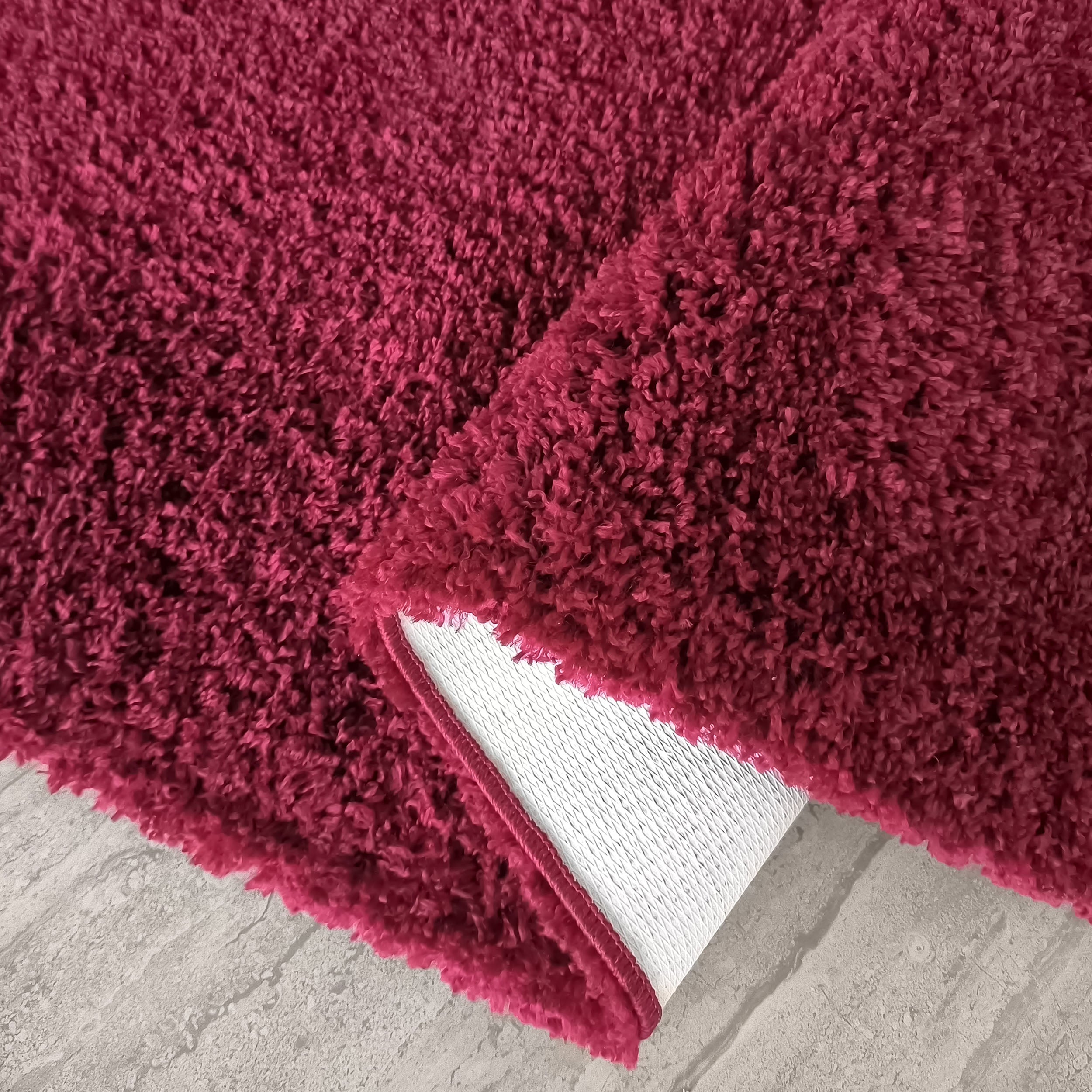 Tapis à poils longs de couleur rouge magenta, partiellement enroulé, laissant apparaître son dessous blanc texturé sur un sol gris clair