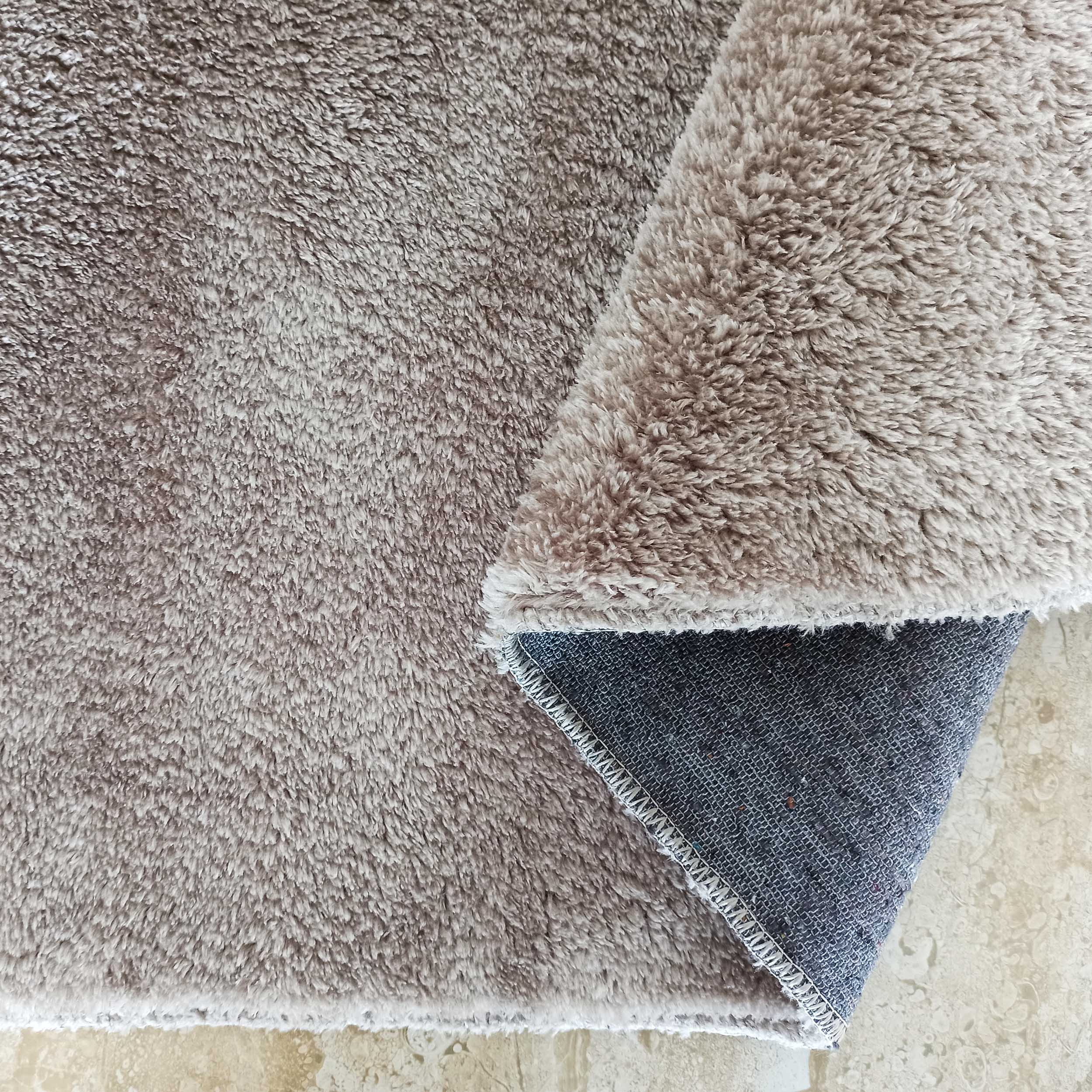 Tapis taupe à poils longs plié, montrant son envers gris quadrillé et la couture blanche sur le sol clair