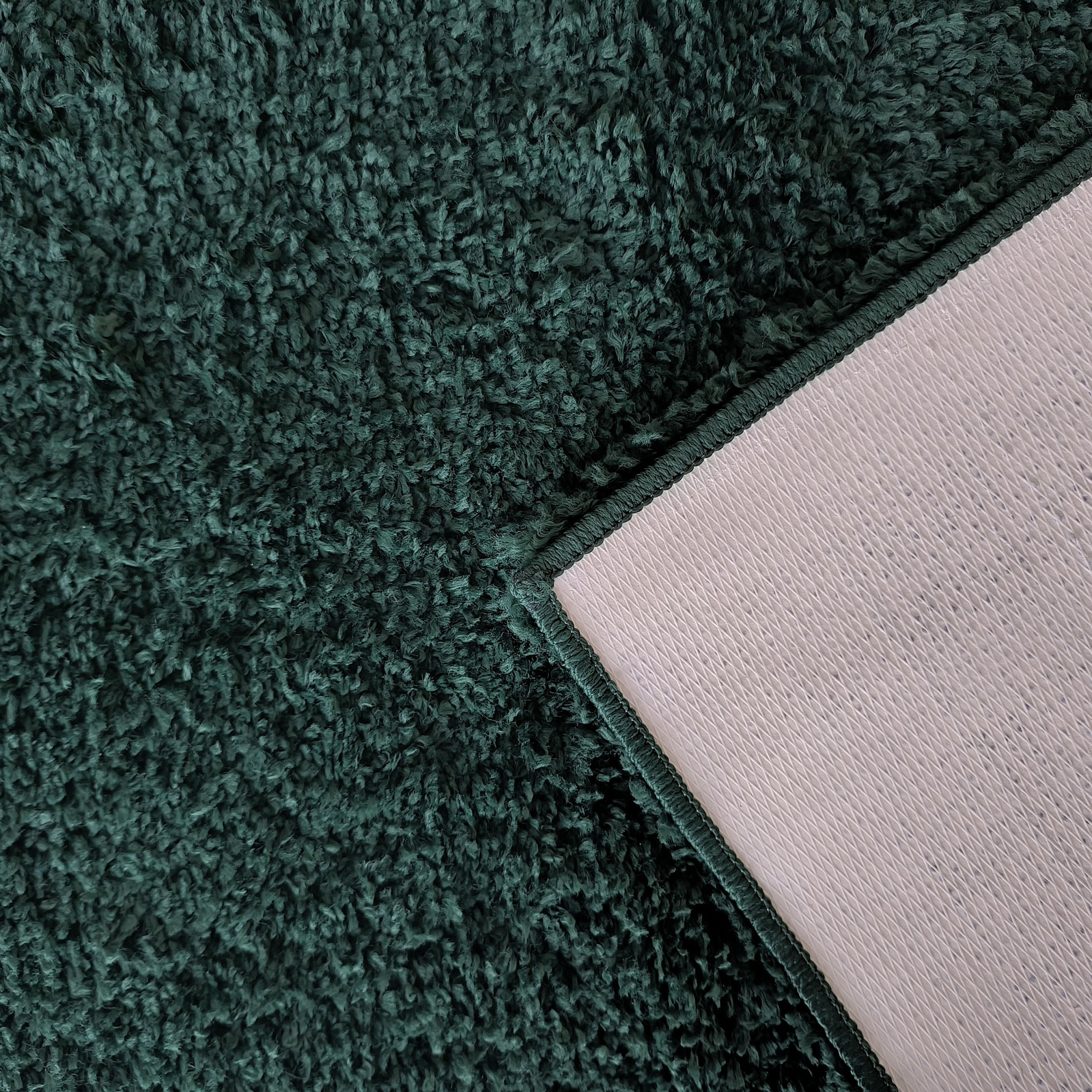 Gros plan sur un tapis shaggy vert foncé, un coin replié dévoile son envers gris clair texturé et sa bordure cousue