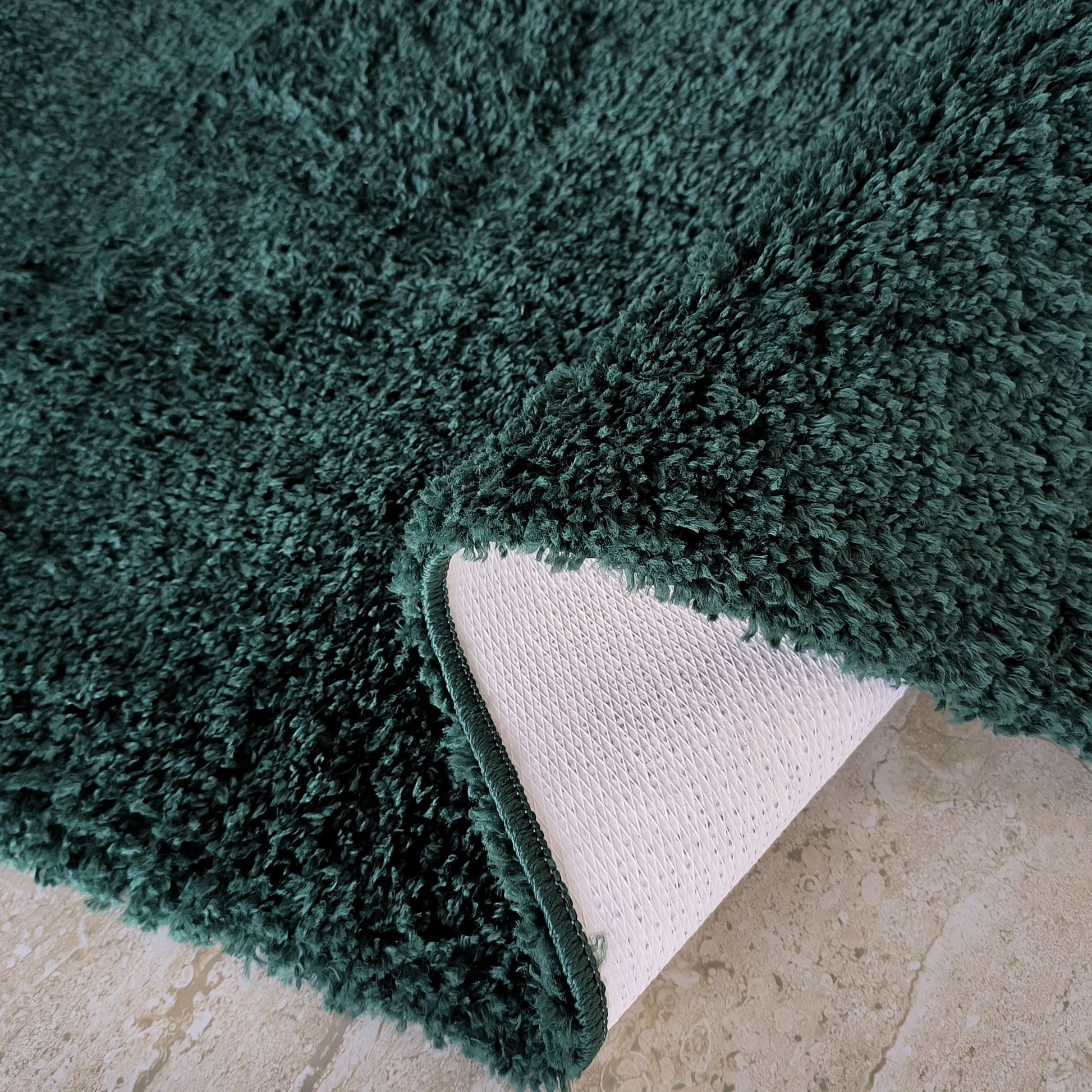 Tapis shaggy vert foncé à longs poils, partiellement replié, révélant son envers blanc quadrillé sur un sol en pierre beige