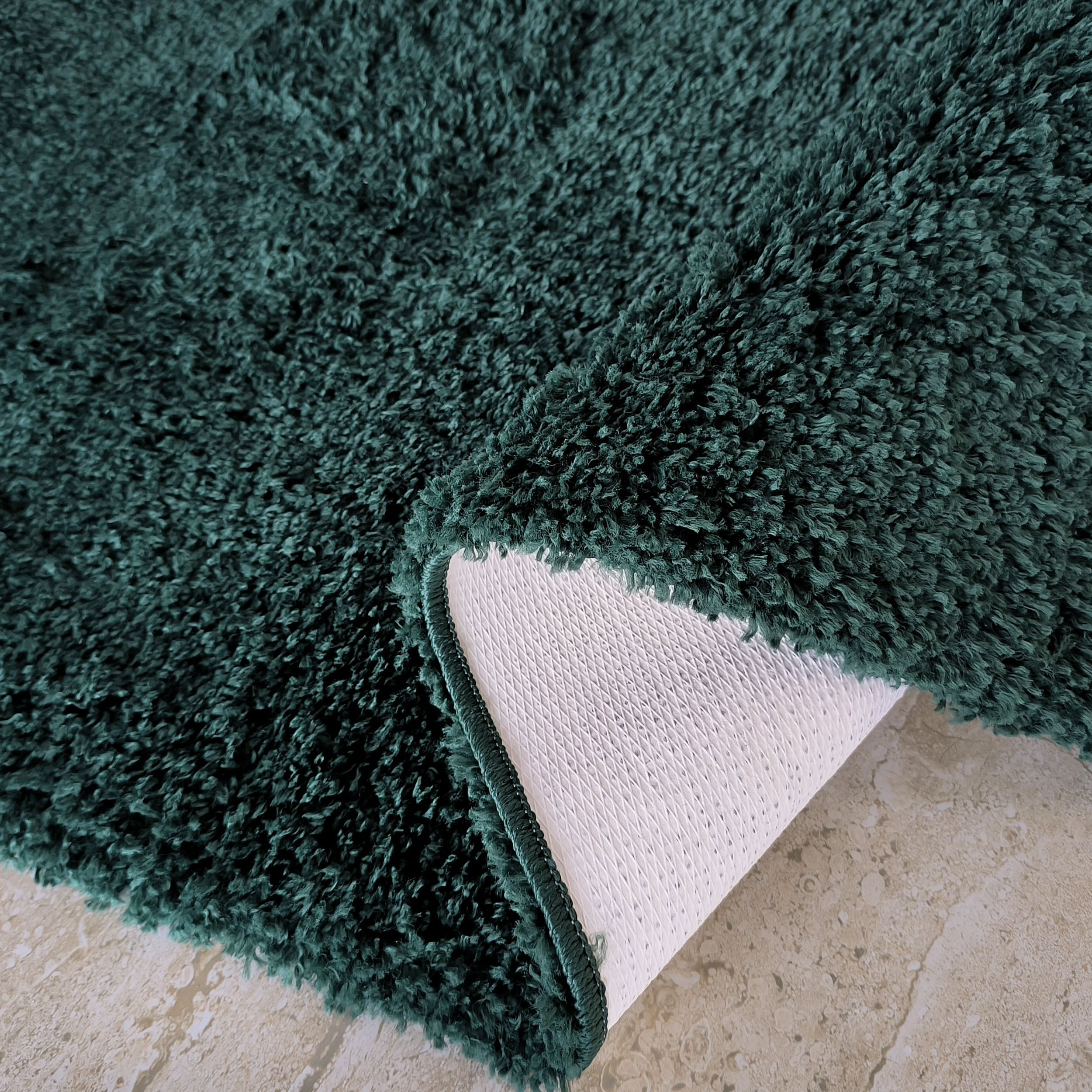 Tapis shaggy vert foncé à longs poils, partiellement replié, révélant son envers blanc quadrillé sur un sol en pierre beige