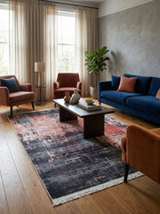 Tapis rectangulaire abstrait aux motifs usés, mêlant des tons de noir, gris foncé, rouille et de légers accents bleus, avec des franges blanches. Il est posé sur un parquet en bois dans un salon décoré