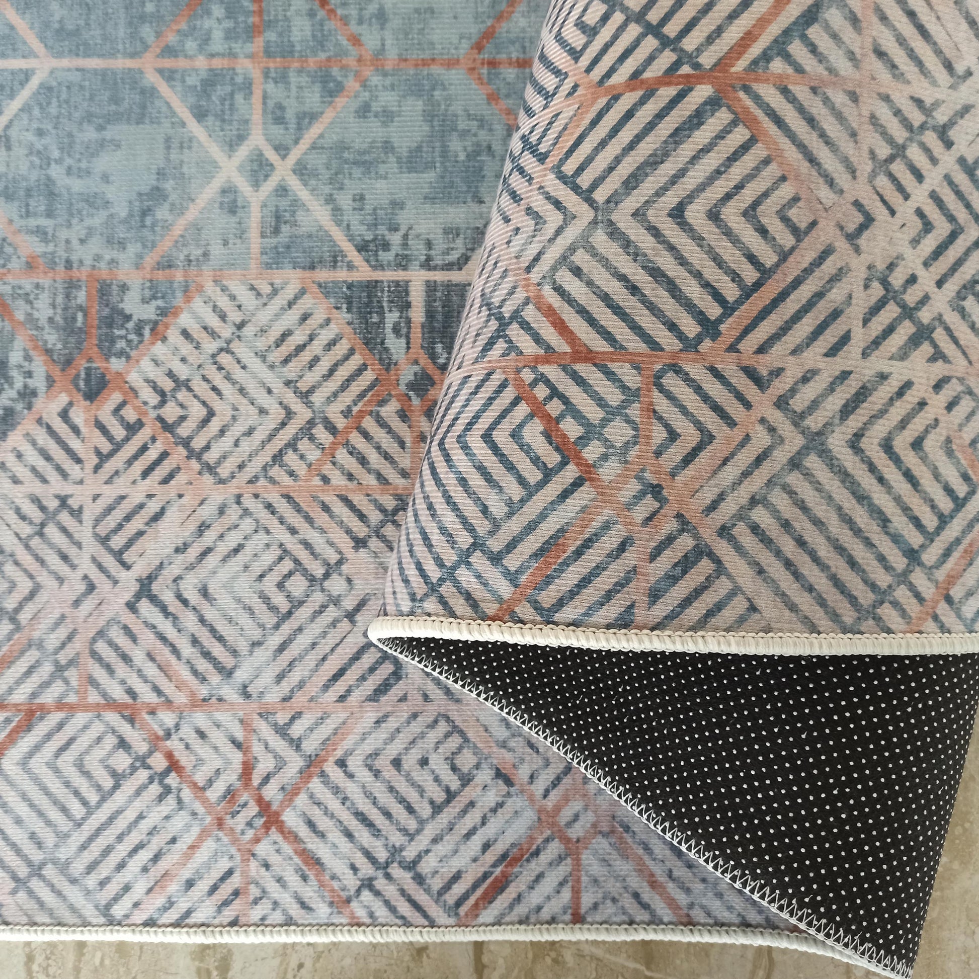 Tapis aux motifs géométriques bleus, beiges et orange rouille, enroulé pour montrer son envers noir à pois blancs et sa bordure blanche