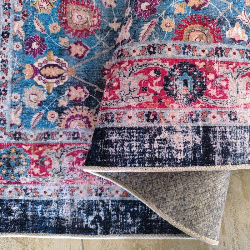 Tapis aux motifs floraux complexes bleu, fuchsia et rouge. Partiellement roulé, il dévoile son envers gris et une bordure foncée