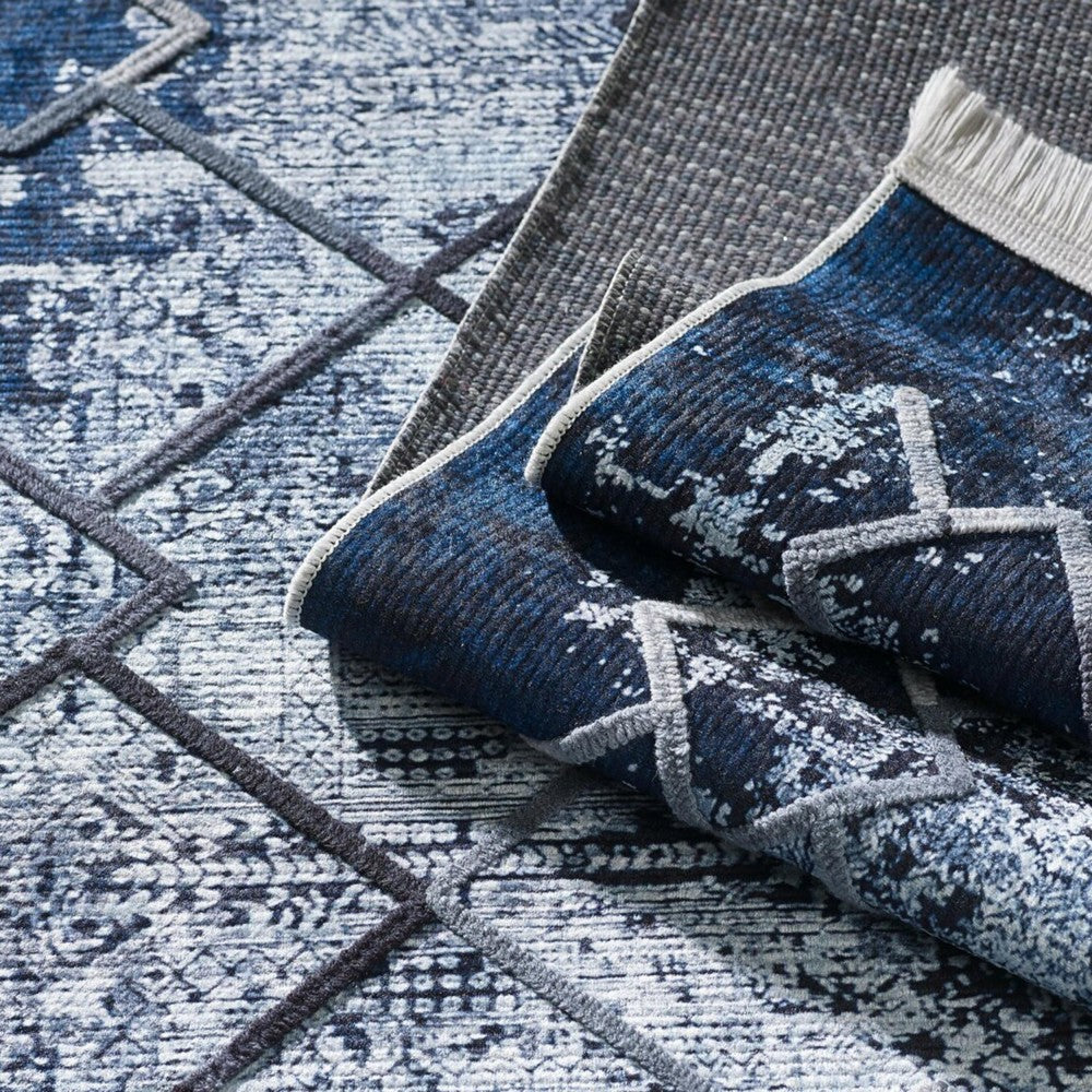 Tapis texturé bleu et blanc aux motifs abstraits et lignes géométriques gris foncé, partiellement roulé sur un tapis gris uni, avec des franges claires visibles