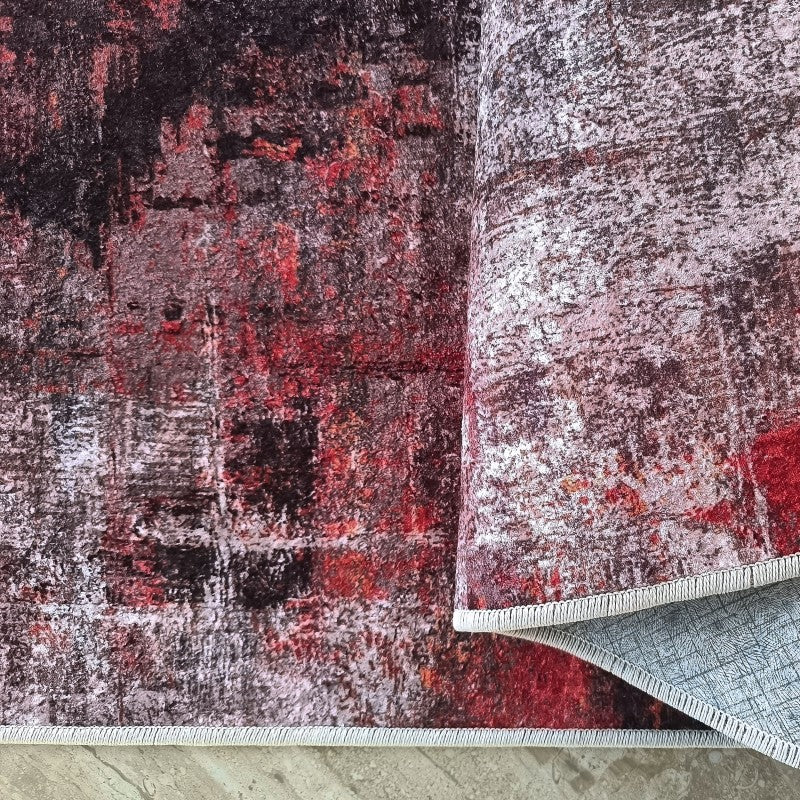 Tapis abstrait aux motifs rouge vif, noir et gris, partiellement roulé, révélant son dessous gris à texture quadrillée