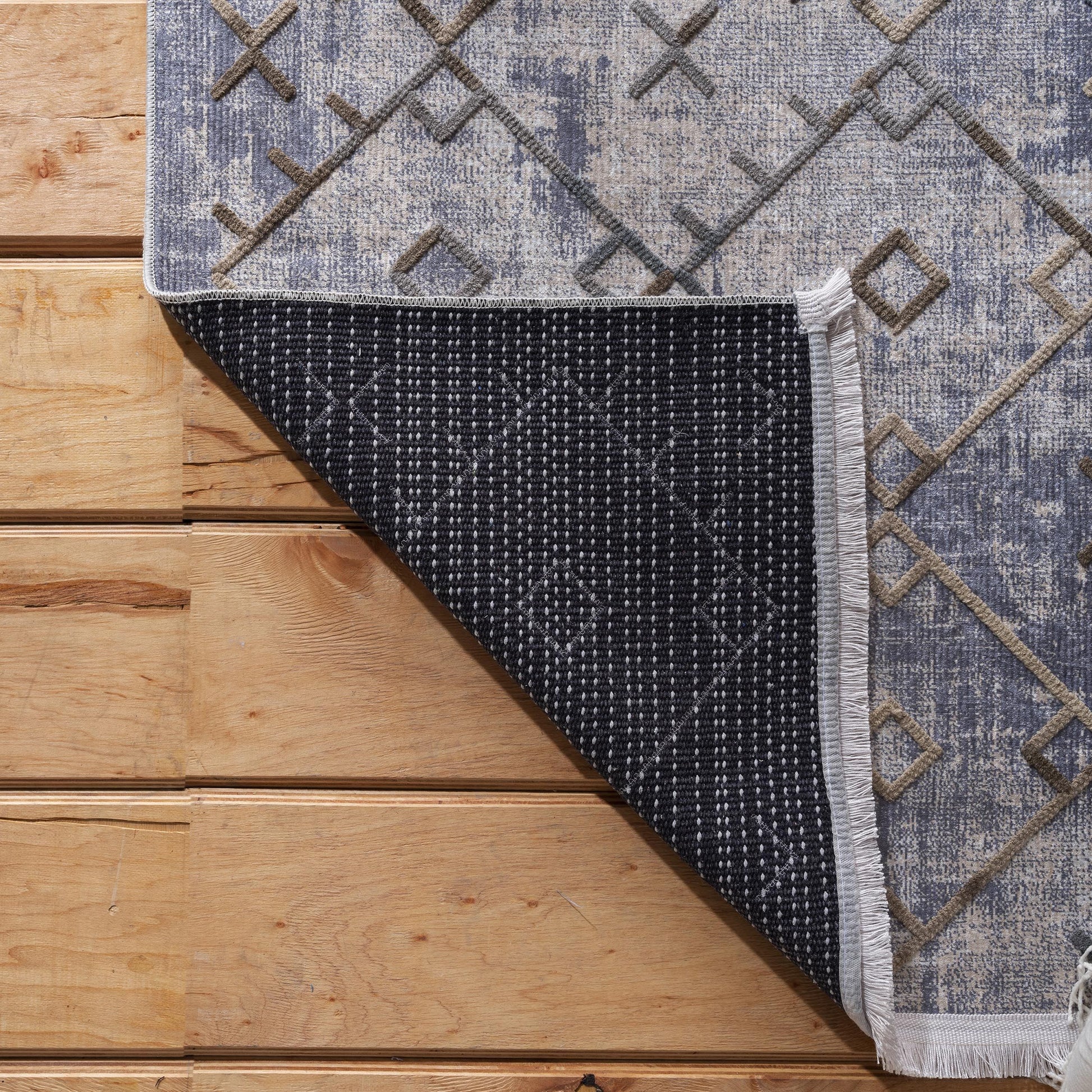 Tapis gris bleu et beige aux motifs géométriques marron en relief, partiellement replié sur un plancher en bois. L'envers noir à petits points blancs et une frange latérale sont visibles