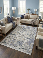 Tapis rectangulaire aux motifs géométriques abstraits gris clair, bleu-gris et taupe sur fond texturé, avec franges, dans un salon contemporain