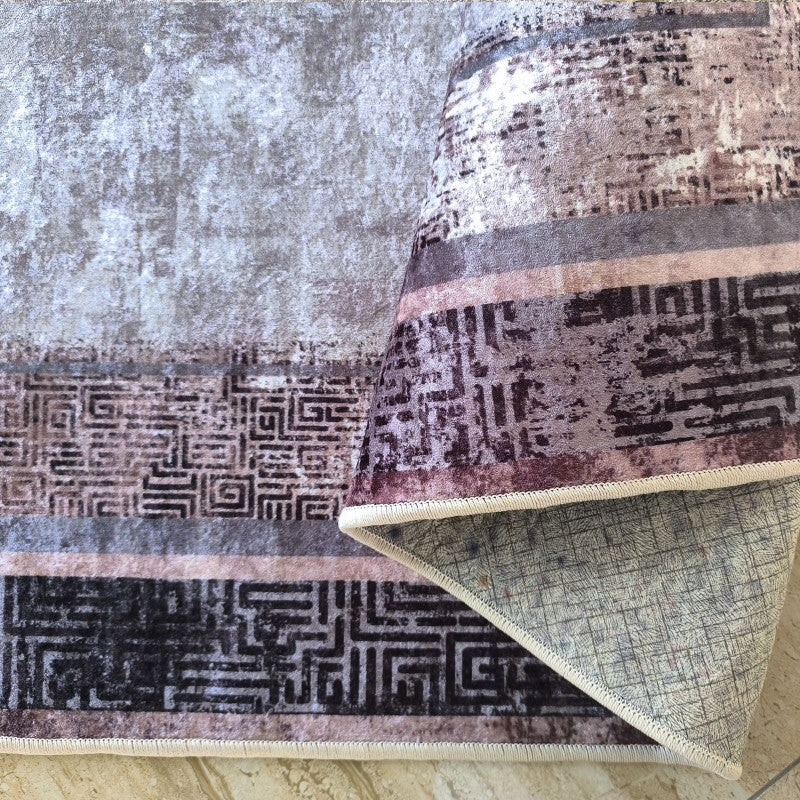 Tapis moelleux aux motifs abstraits et géométriques dans des tons de gris, marron et rose. Un coin est replié, révélant son dos texturé