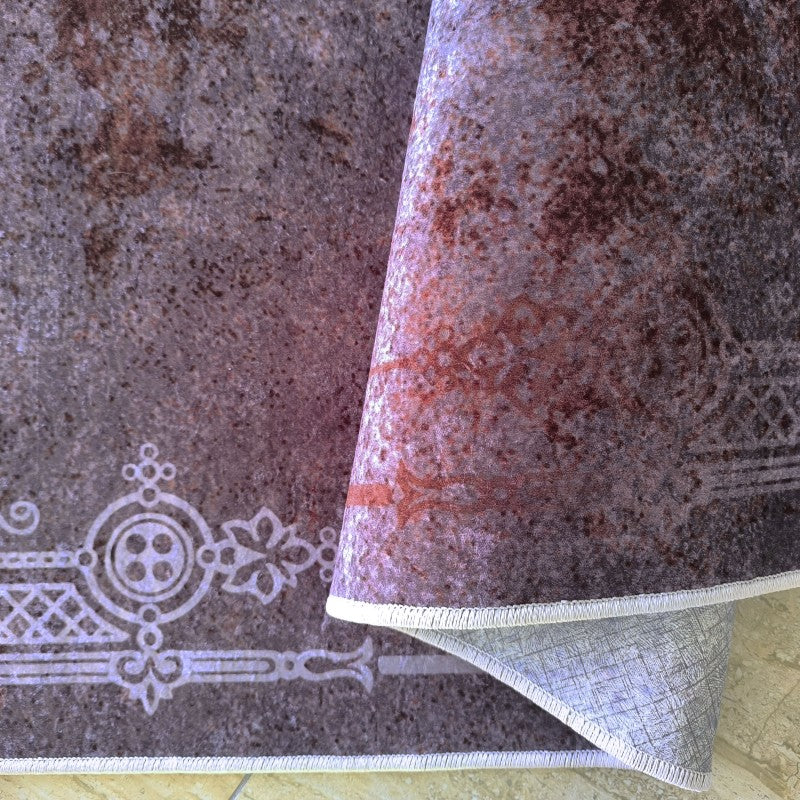 Tapis doux aux teintes de mauve, marron et gris avec un motif texturé et une bordure ornementale claire, un coin est plié