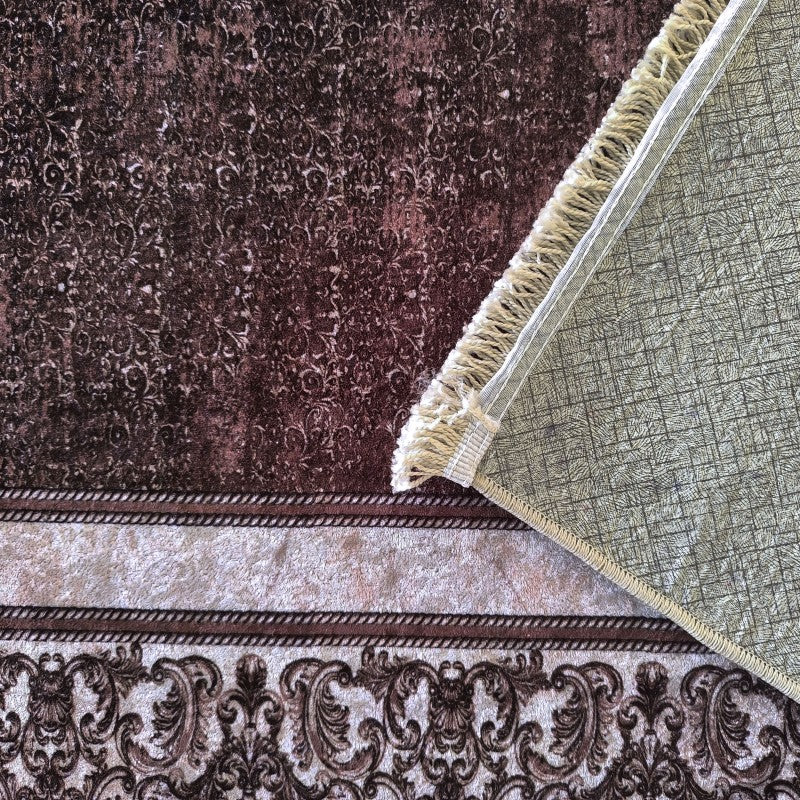 Tapis en velours marron foncé à motifs baroques délavés, bordure beige aux motifs similaires, un coin relevé montrant le dessous vert texturé et des franges crème
