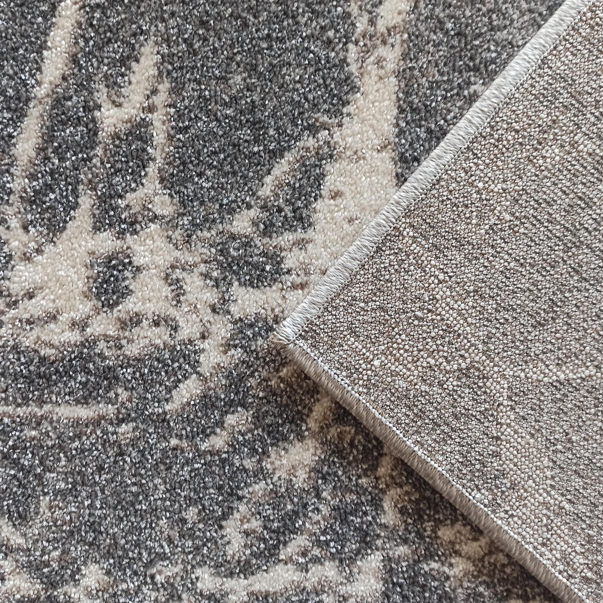 Tapis texturé aux motifs abstraits gris foncé et crème, un coin soulevé montre son envers tissé et sa bordure cousue